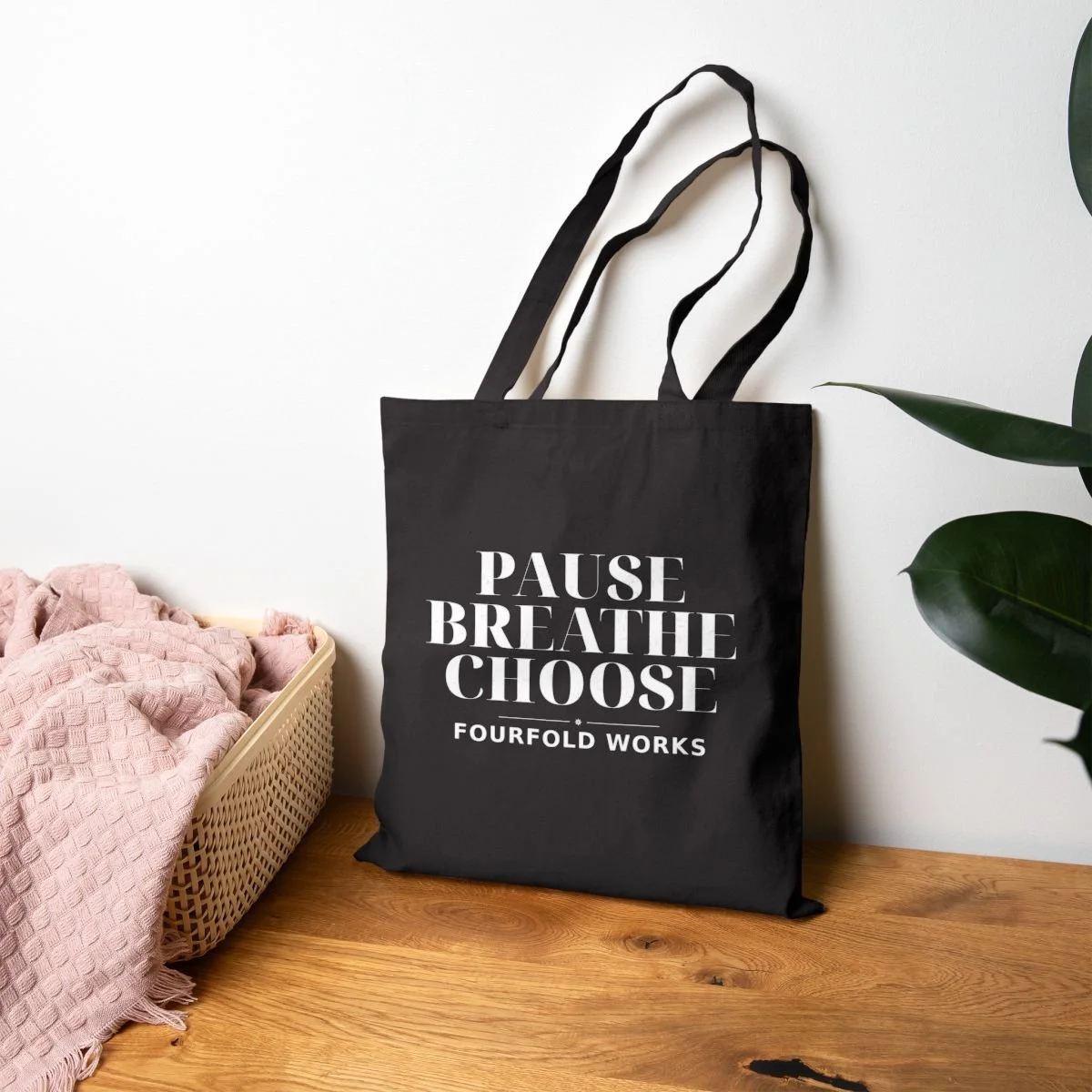 Black Pause Breathe Choose tote hanging above wood beside a wicker basket, pink throw, and green leaves.