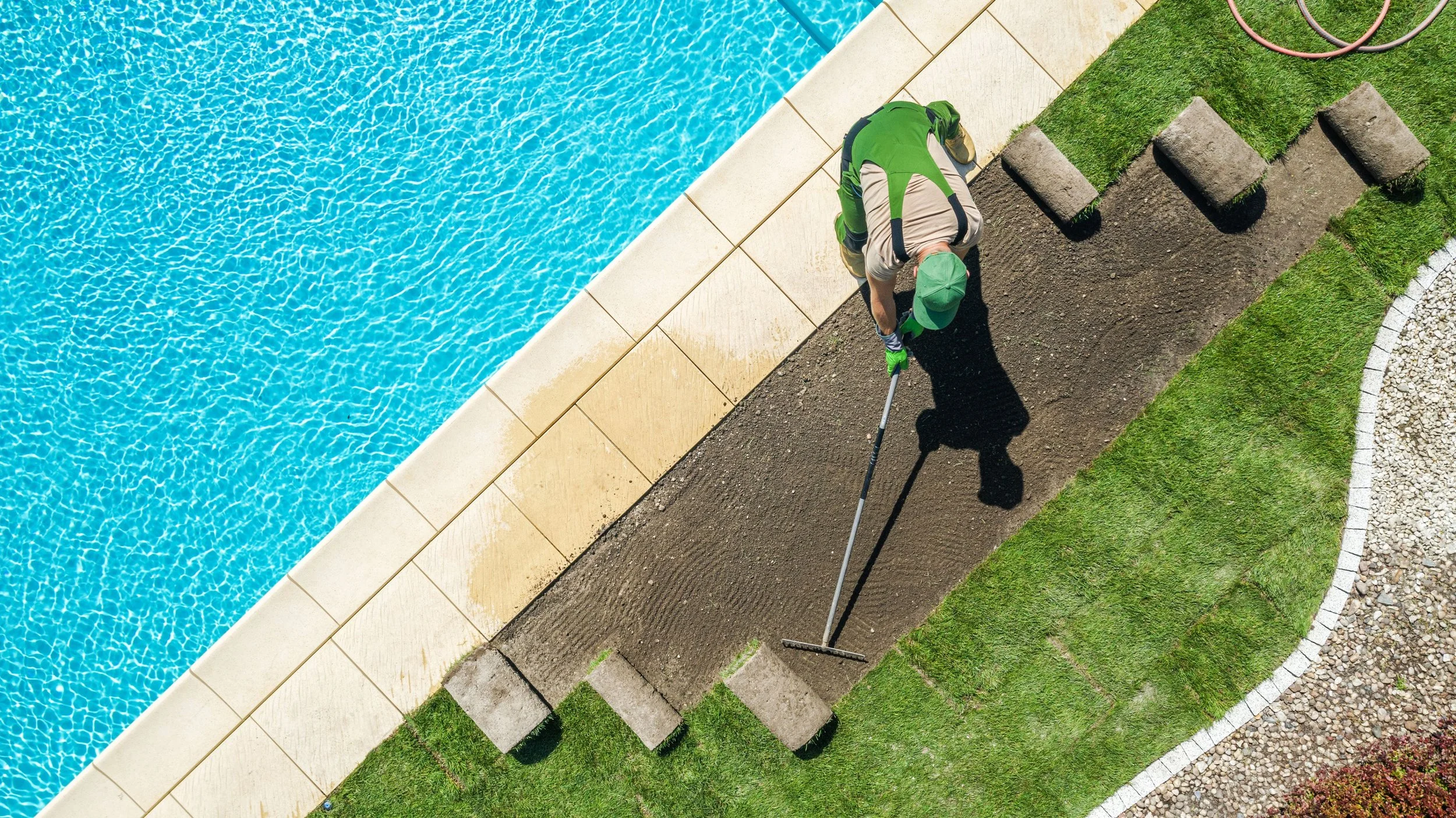 A person in a green cap and gloves is planting grass sod along a garden edge near a swimming pool. The pool water is blue and clear, and there are stepping stones and grass patches in the garden.