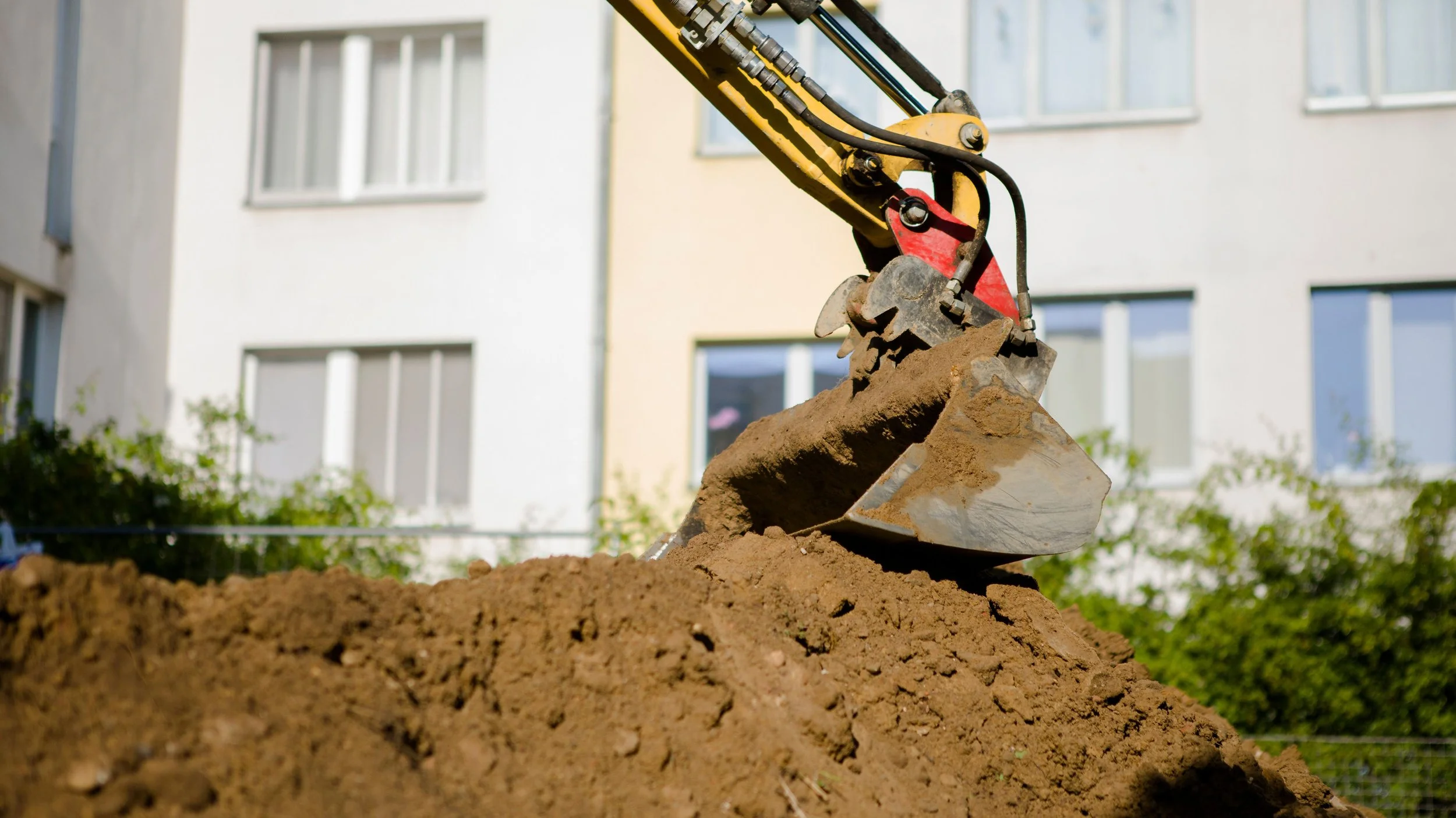 Close-up of an excavator bucket filled with dirt at a construction site, with a residential building with multiple windows in the background.