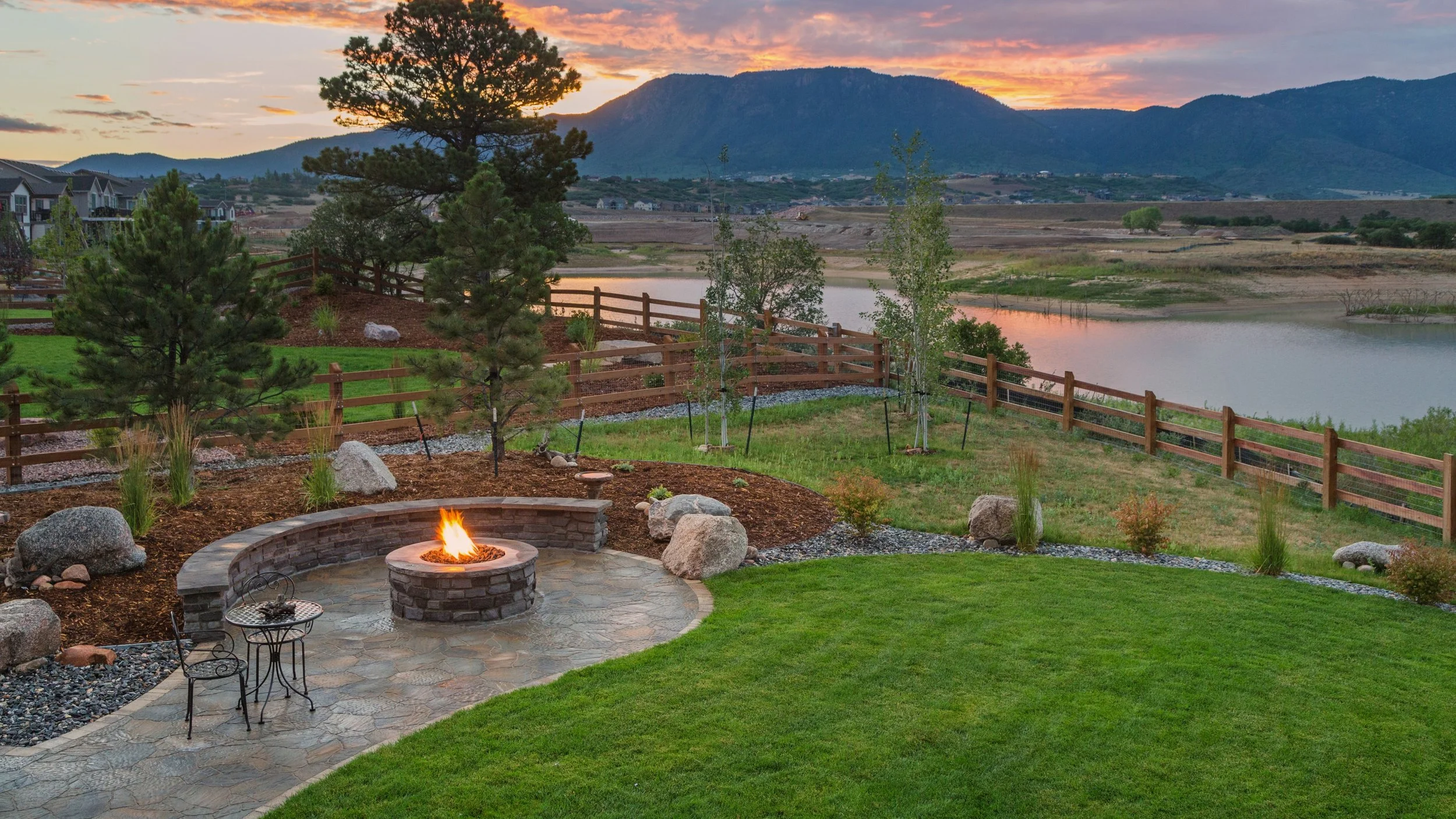 A backyard patio with a fire pit, two chairs, and a small table, overlooking a lake and mountains during sunset. The yard has a well-maintained lawn, trees, and a wooden fence.