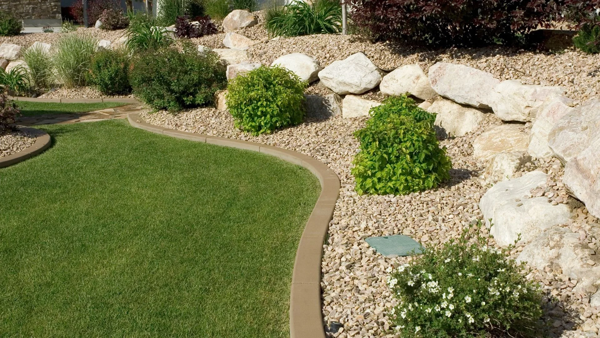 A landscaped garden with a green lawn, a curved border, and a rocky area with various plants and shrubs.