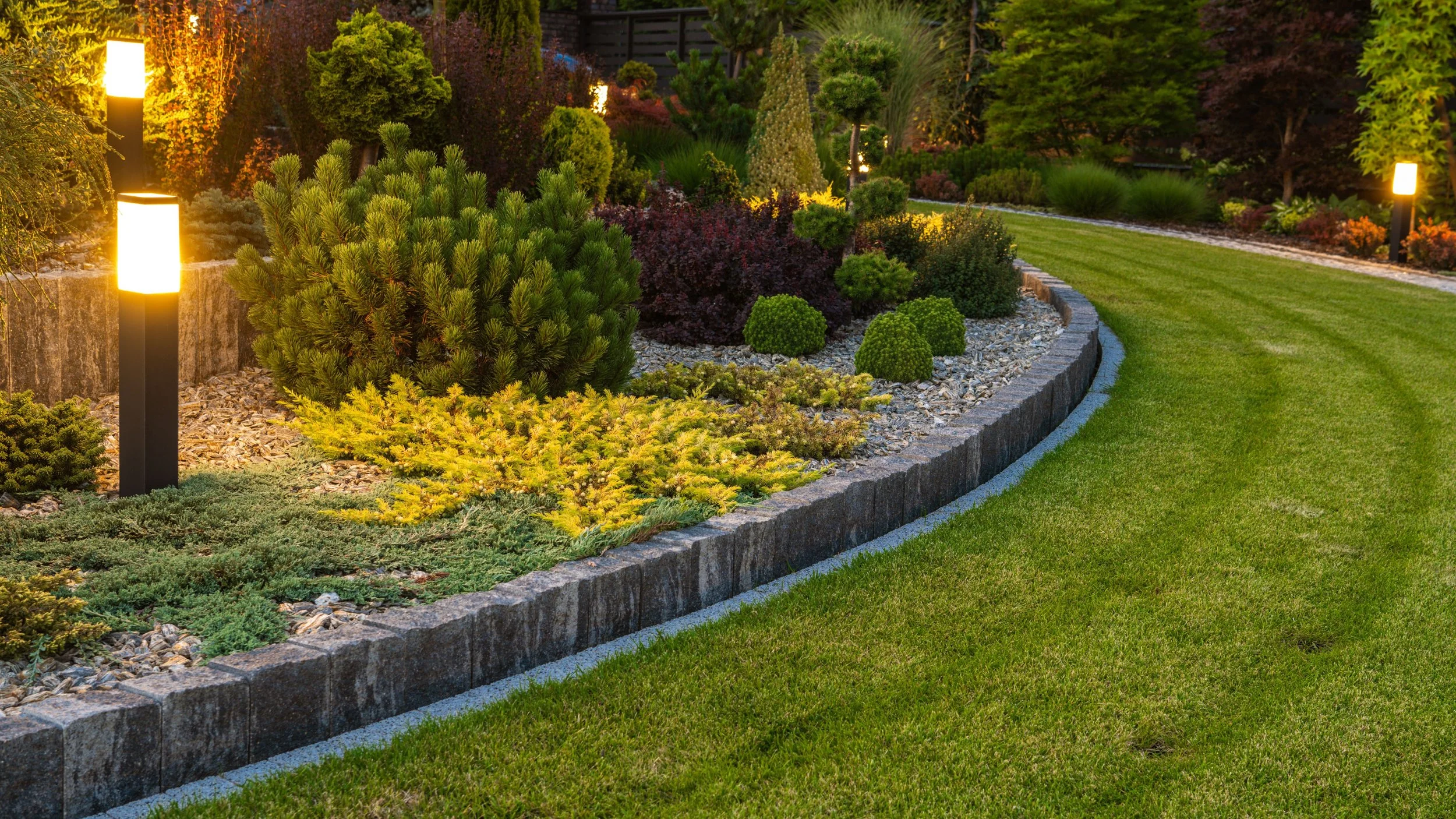 A landscaped backyard garden at dusk with a curved brick or stone border, various green shrubs and plants, and several modern fence post lights illuminating the area.