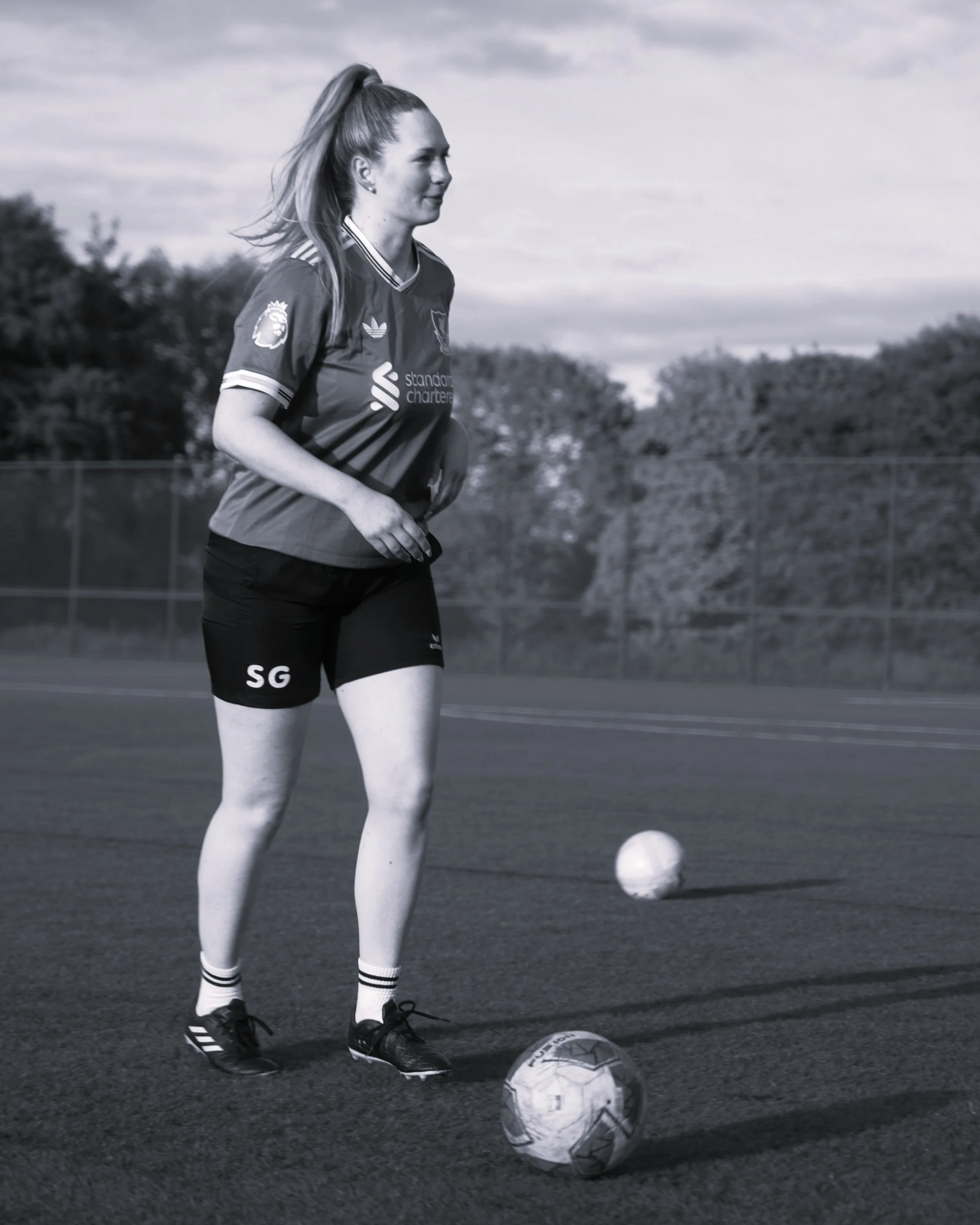 A female football player on a field preparing to kick a ball, wearing a football shirt and shorts.