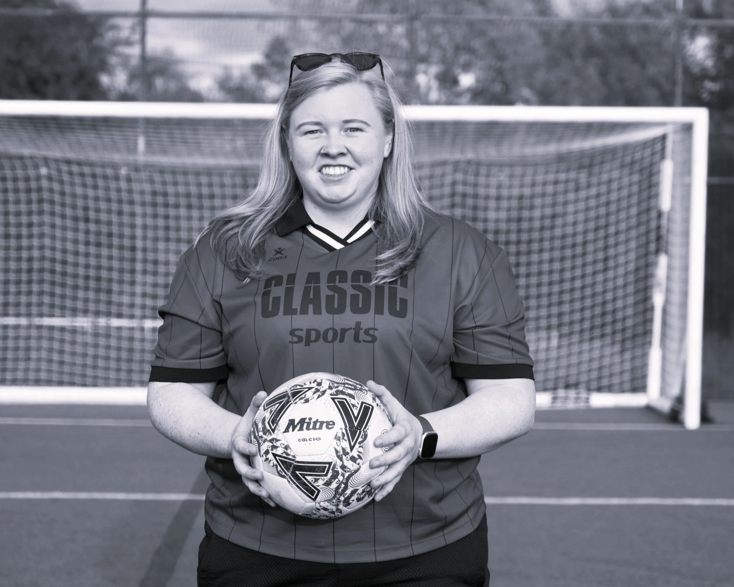 A woman in sports gear standing in front of a football goal, holding a soccer ball with both hands, outdoors on a cloudy day.