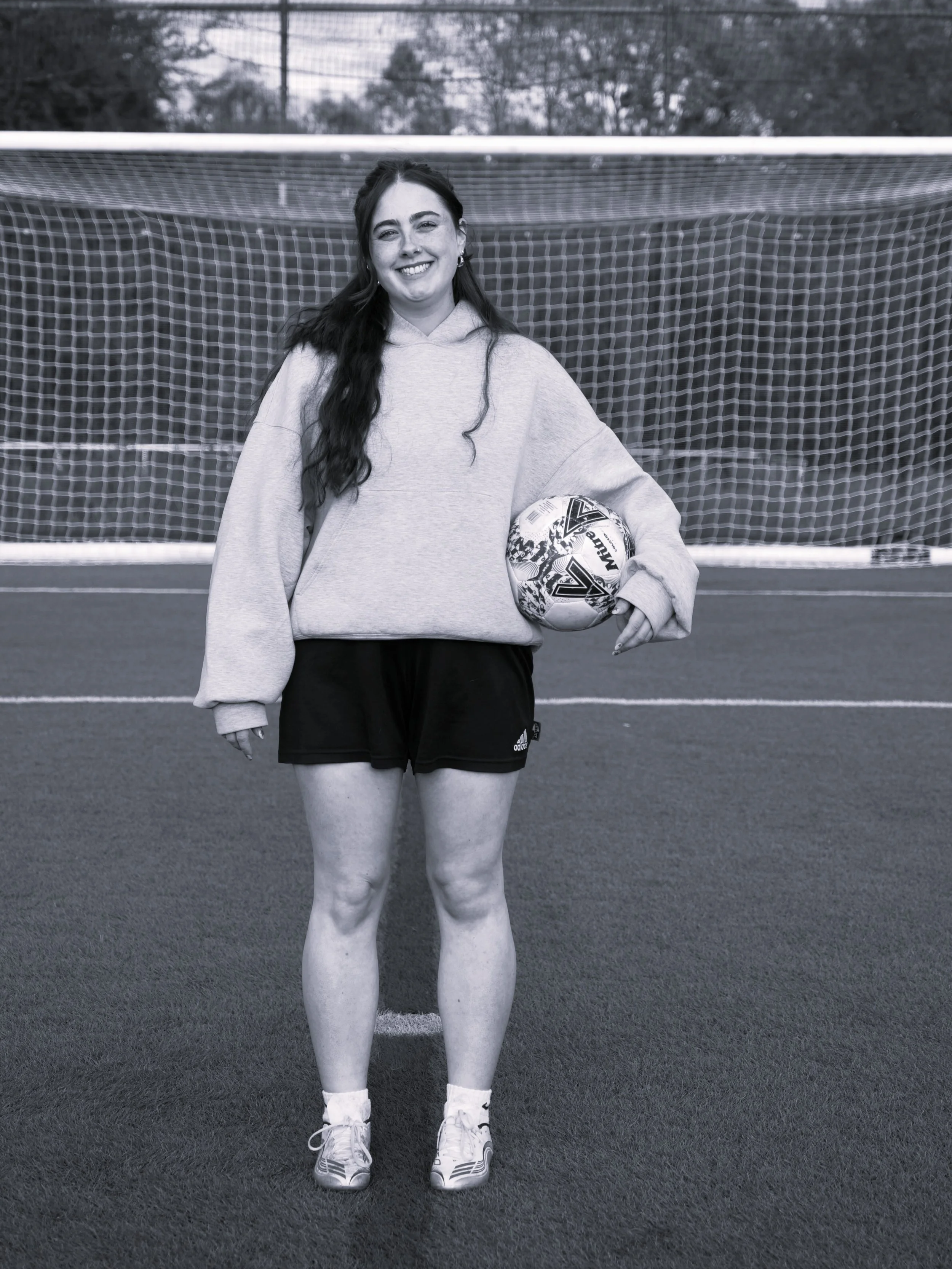 A young woman standing on a football field holding a soccer ball, smiling, in front of a goal net.