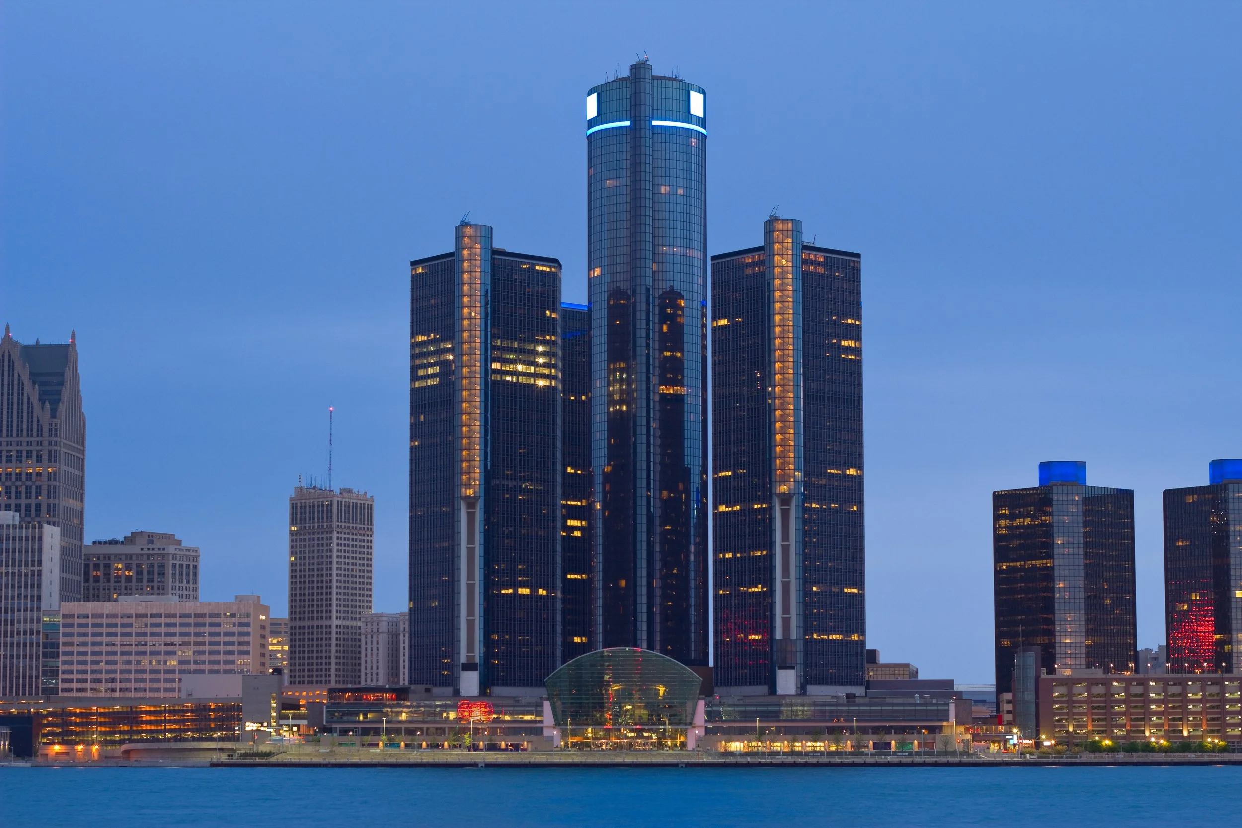 Night cityscape featuring tall glass skyscrapers with illuminated windows and blue accent lighting, city skyline reflected in water in foreground.