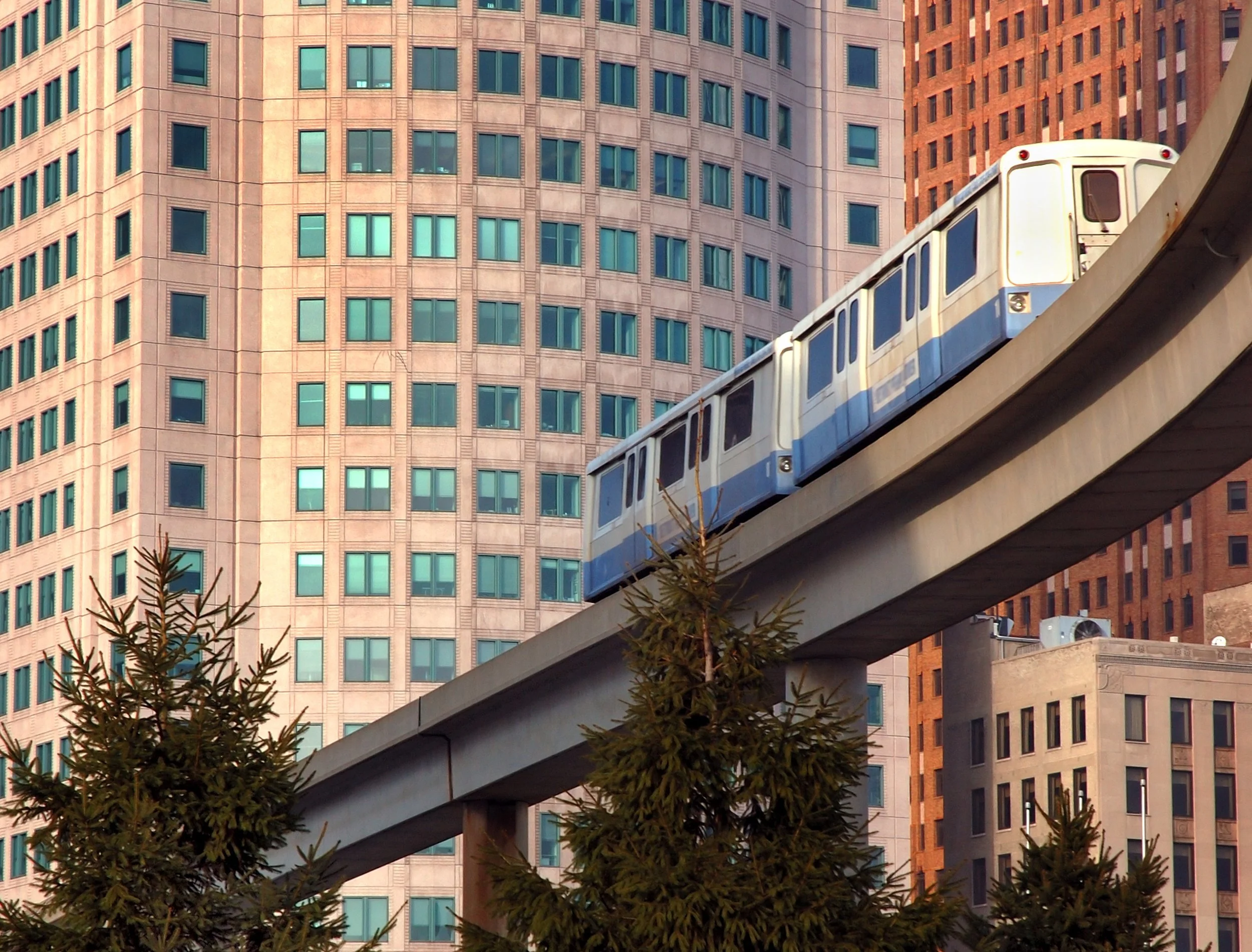 A train moving on an elevated track with a backdrop of tall city buildings and some trees.