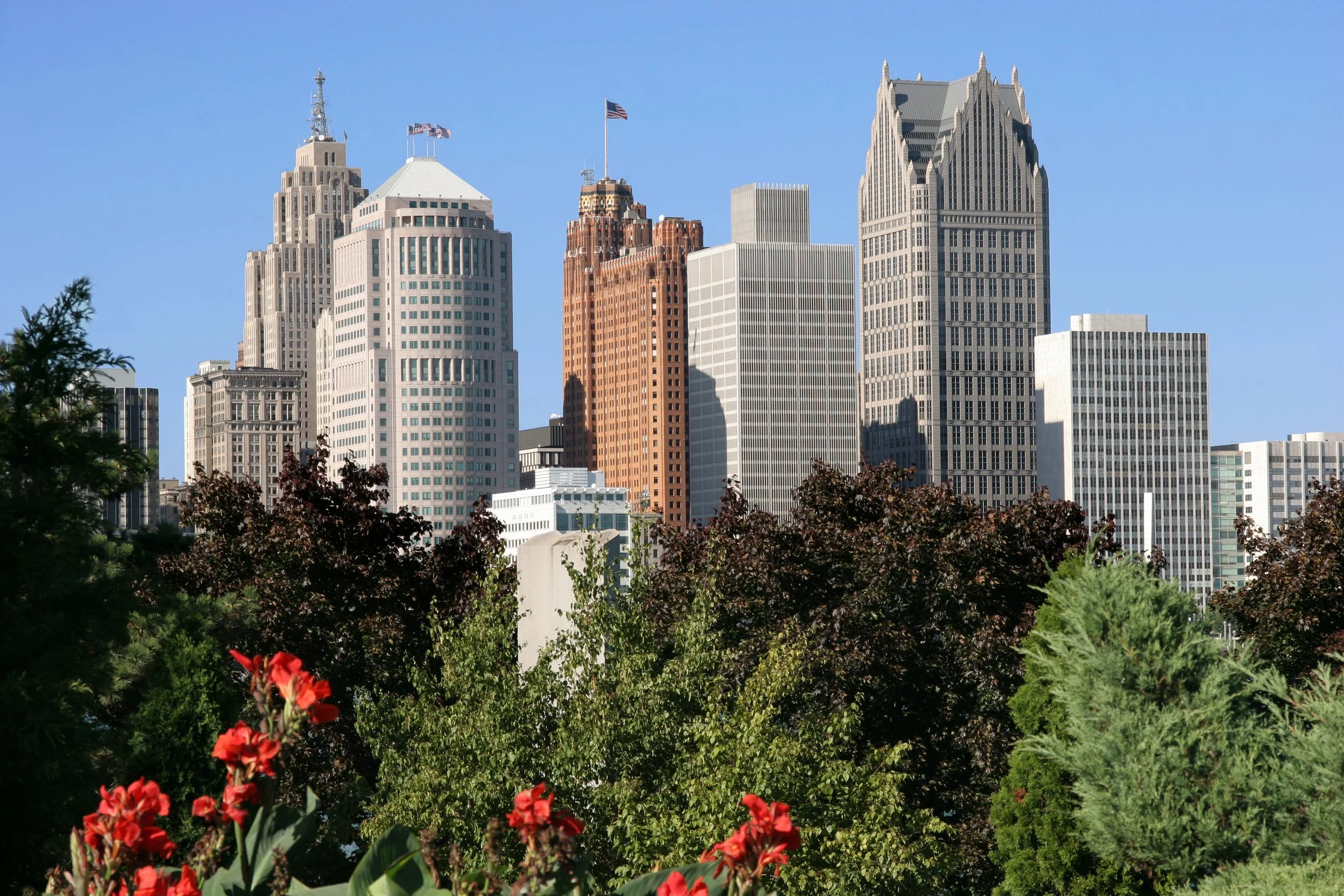 View of downtown skyscrapers framed by trees and red flowers in the foreground under a clear blue sky.
