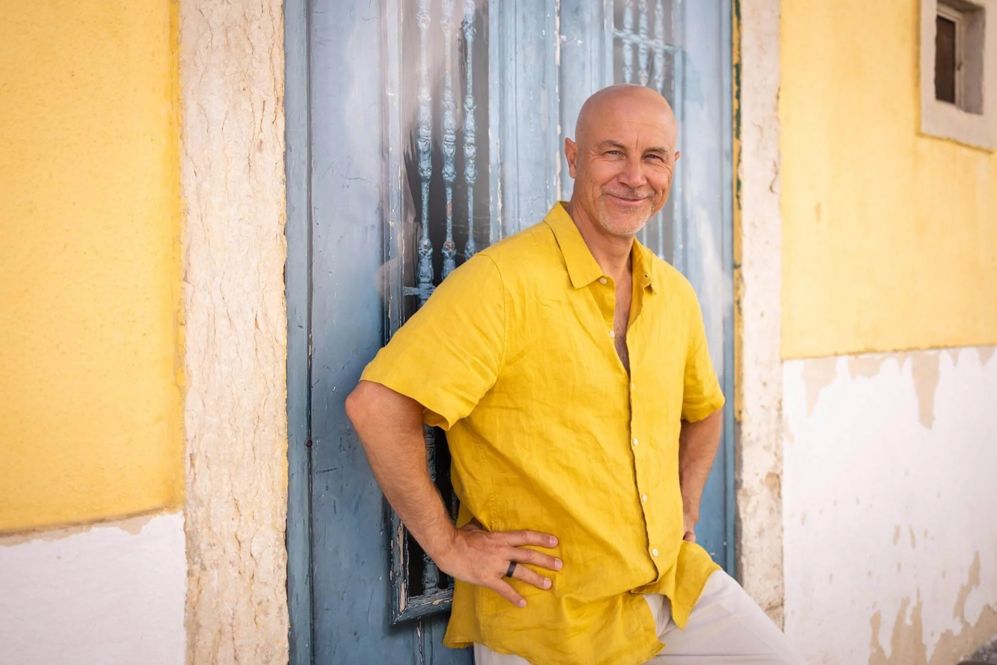 A middle-aged man with a shaved head and a goatee, smiling, standing against a vintage blue wooden door. He is wearing a bright yellow short-sleeved button-up shirt and white pants, with his hands on his hips.