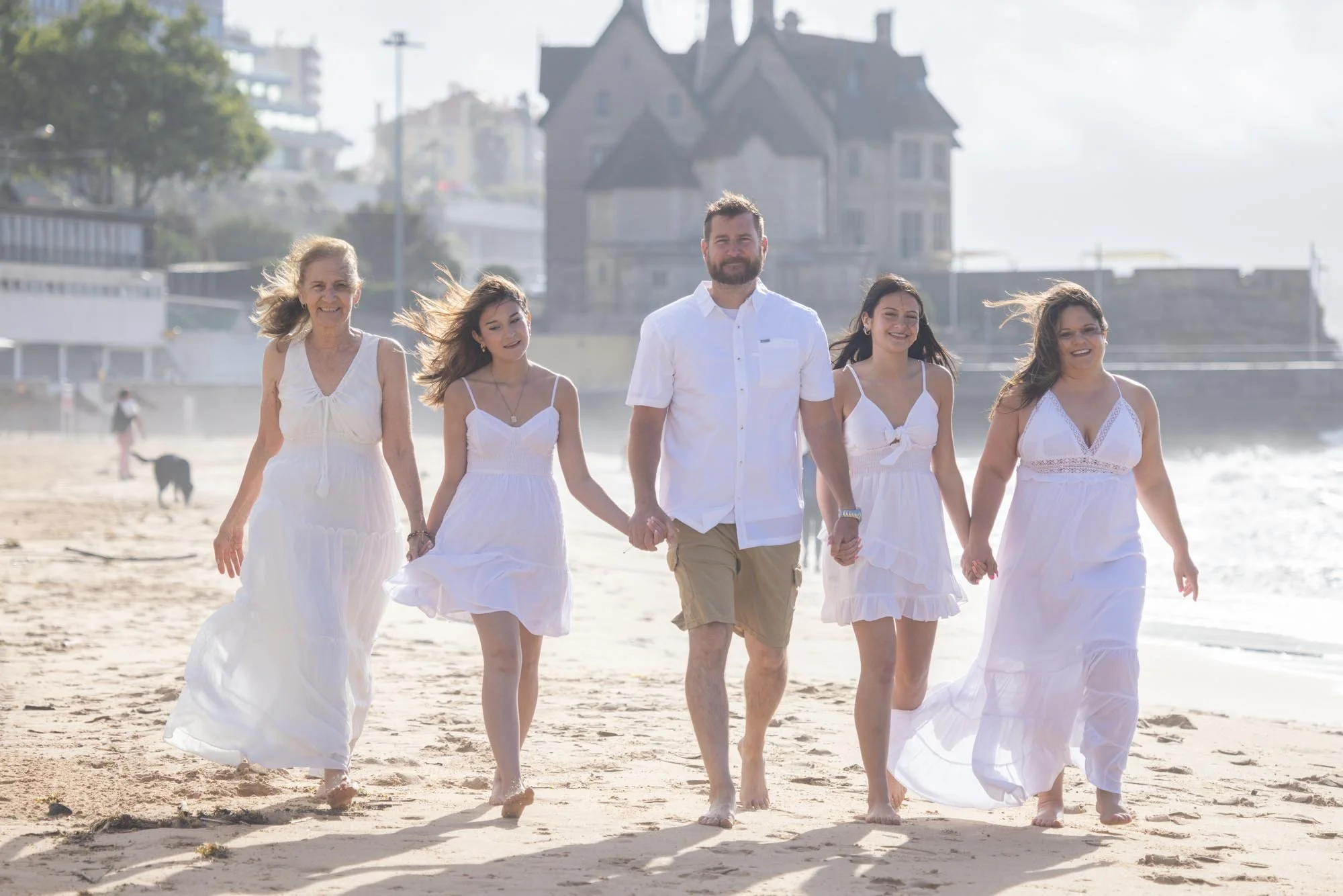 People walking on the beach, holding hands, dressed in white, with a large house and city buildings in the background.