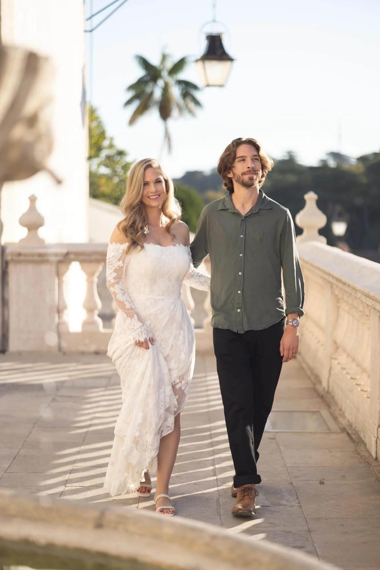 A couple is walking outdoors on a sunny day, with the woman wearing a white lace wedding dress and the man in casual dark pants and a green shirt.