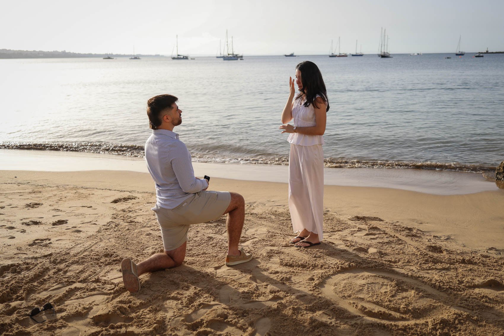 A man is proposing marriage to a woman on the beach during sunset, with sailboats in the background. Praia da Rainha beach in Cascais.