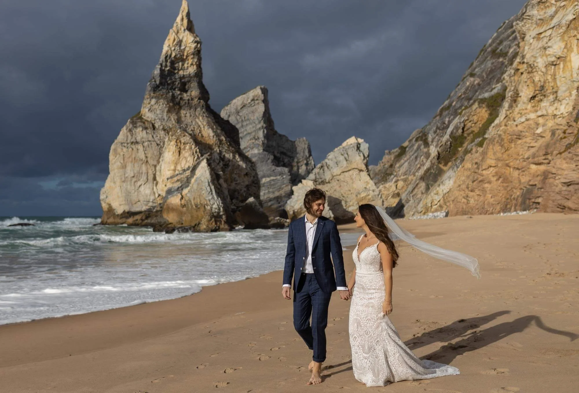 A bride and groom walking barefoot on a sandy beach, holding hands, with large rock formations in the background and a cloudy sky overhead.