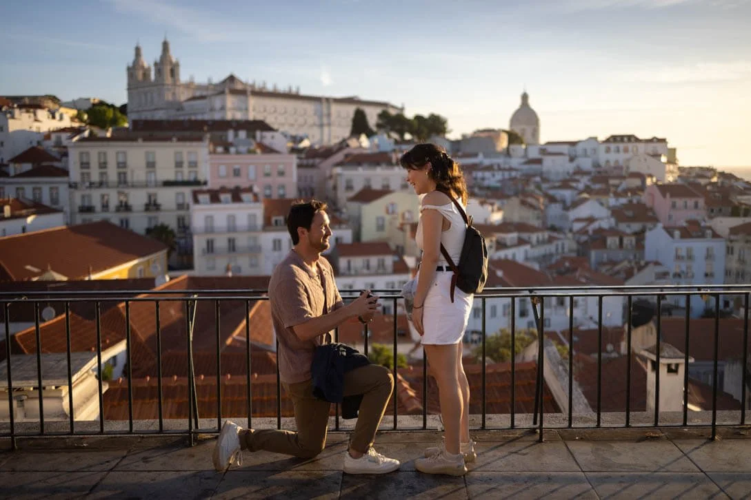 A man proposing to a woman on a rooftop with a cityscape in the background at sunset.