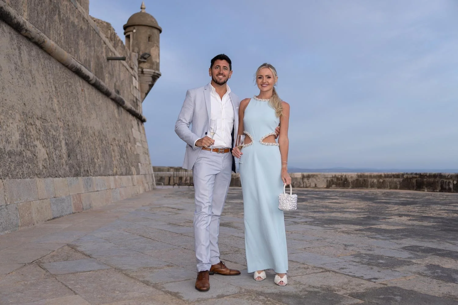 A man and woman in elegant attire standing outside on a stone-paved area near a large stone wall and fortress tower, holding champagne glasses. The woman is wearing a light blue dress with cutouts and holding a small beaded handbag. The man is dresse