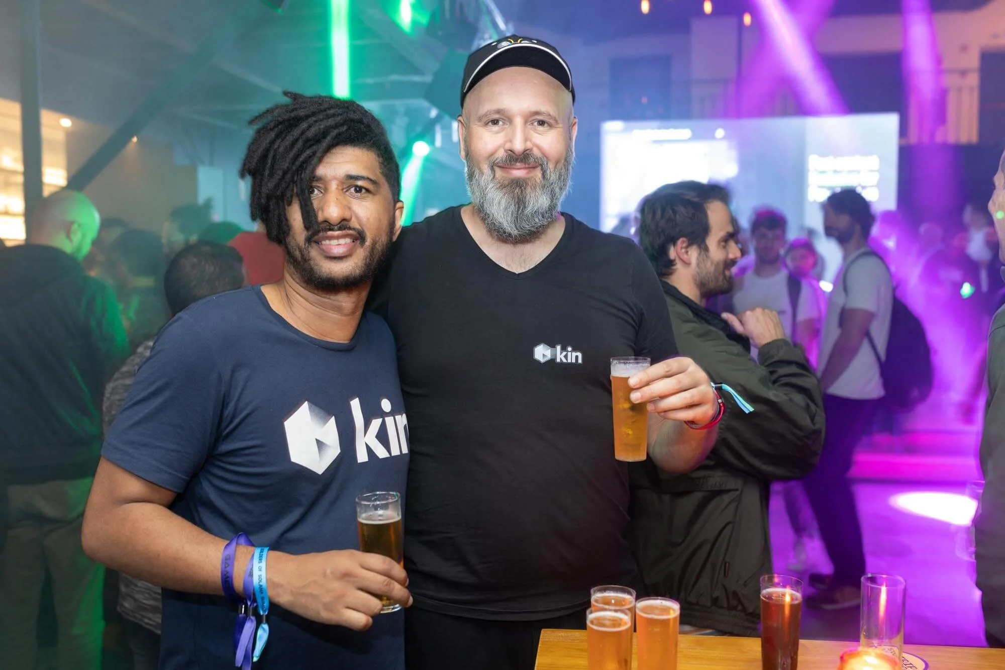 Two men smiling in a lively, colorful party or club setting, holding beers, with people dancing and lights in the background.