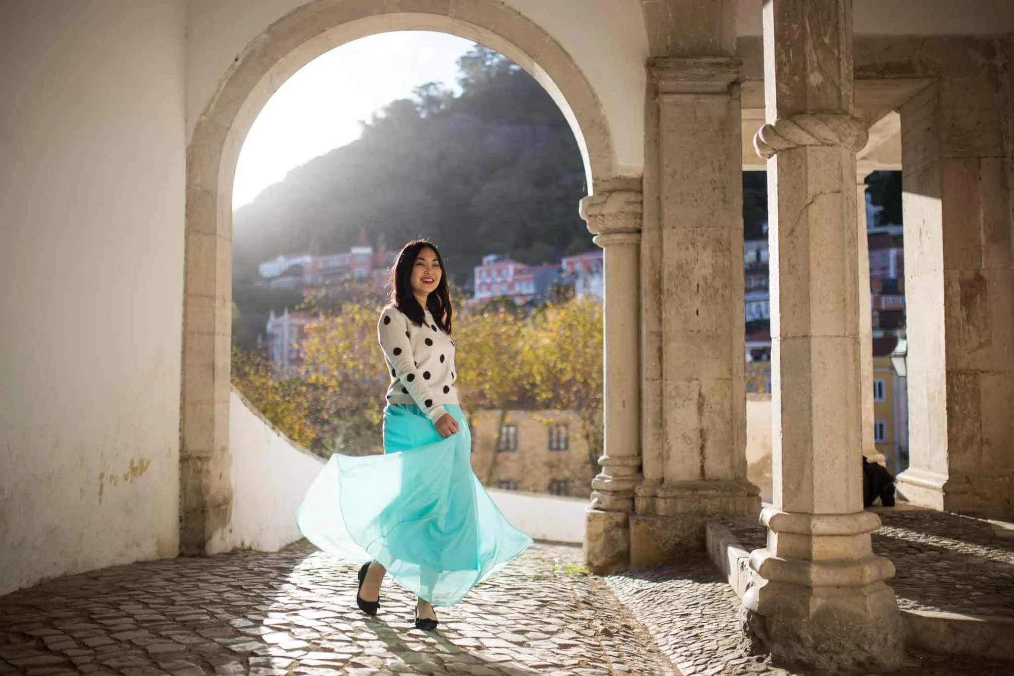 A woman smiling and twirling in a turquoise skirt under an arched stone structure with a scenic hillside and buildings in the background.