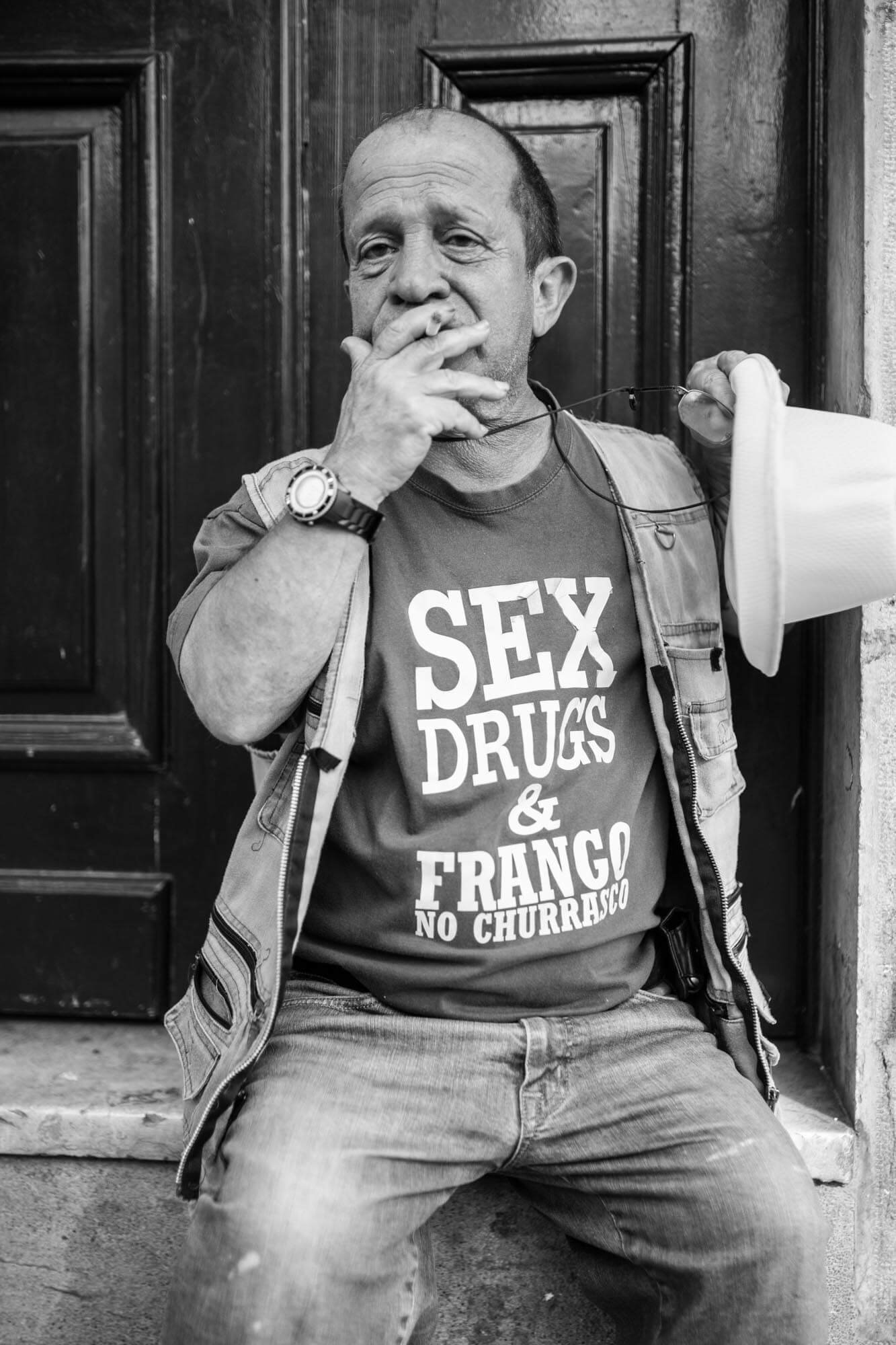 A man is sitting on a step in front of a dark wooden door, holding a megaphone, touching his lips with one hand, and wearing a t-shirt that says 'SEX, DRUGS, & FRANGO NO CHURRASCO' in Portuguese. The image is in black and white.