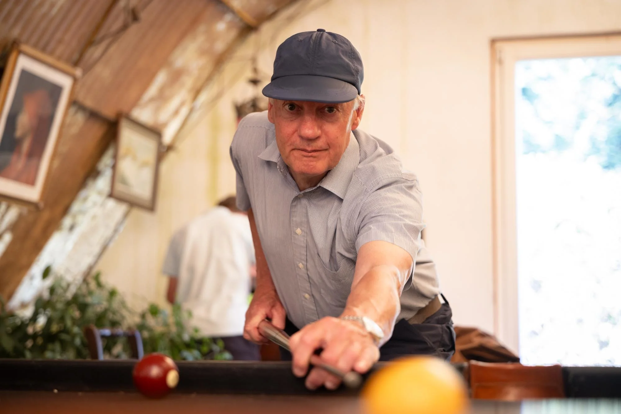 An elderly man in a gray cap and light gray button-up shirt playing pool indoors, leaning over the table, aiming to hit the yellow ball, with a red ball nearby. The room has wood-paneled walls and framed pictures, and a window letting in natural ligh
