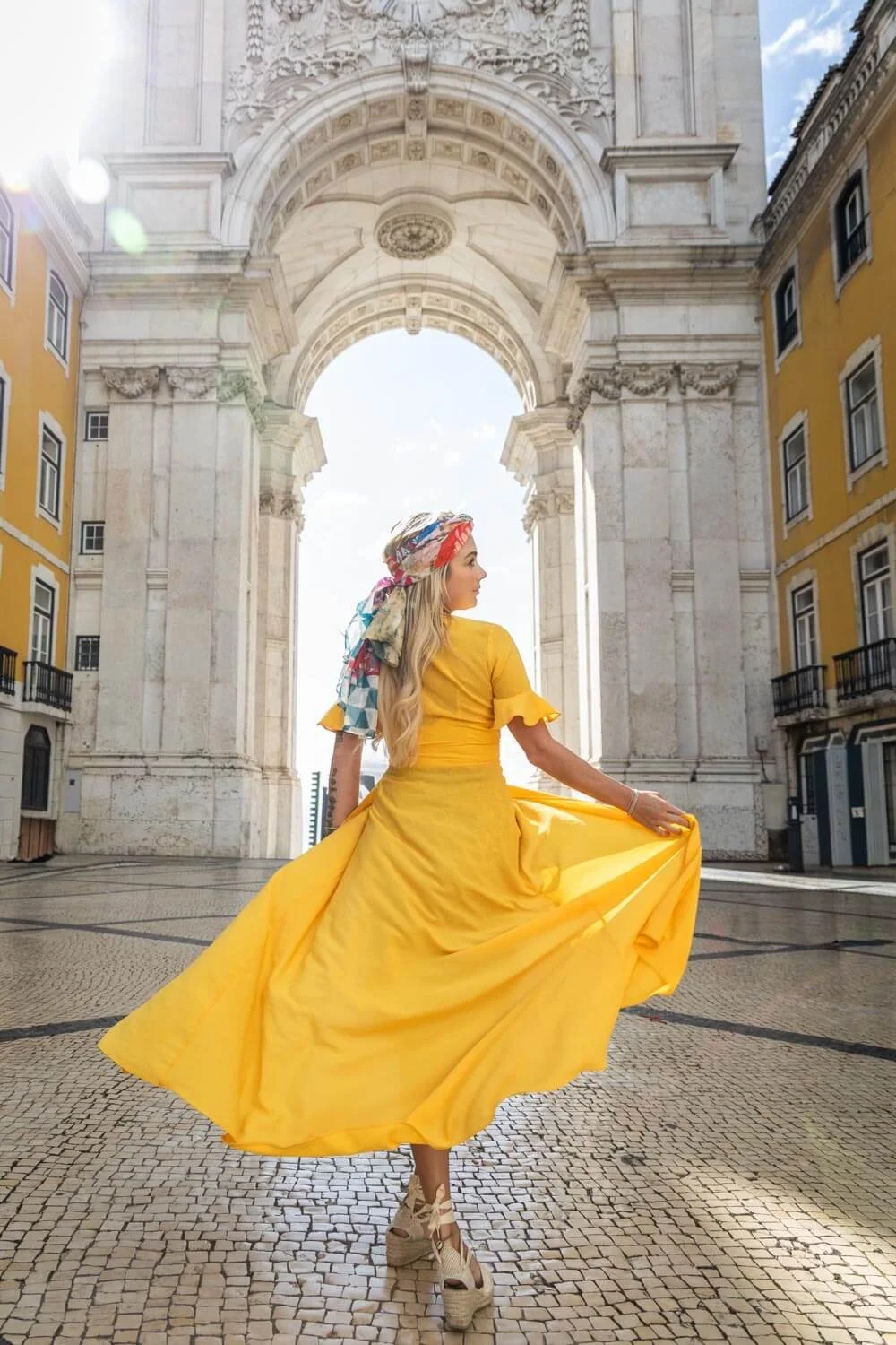 A woman in a flowing yellow dress and patterned headscarf stands in front of an ornate stone archway, with buildings and cloudy sky in the background.