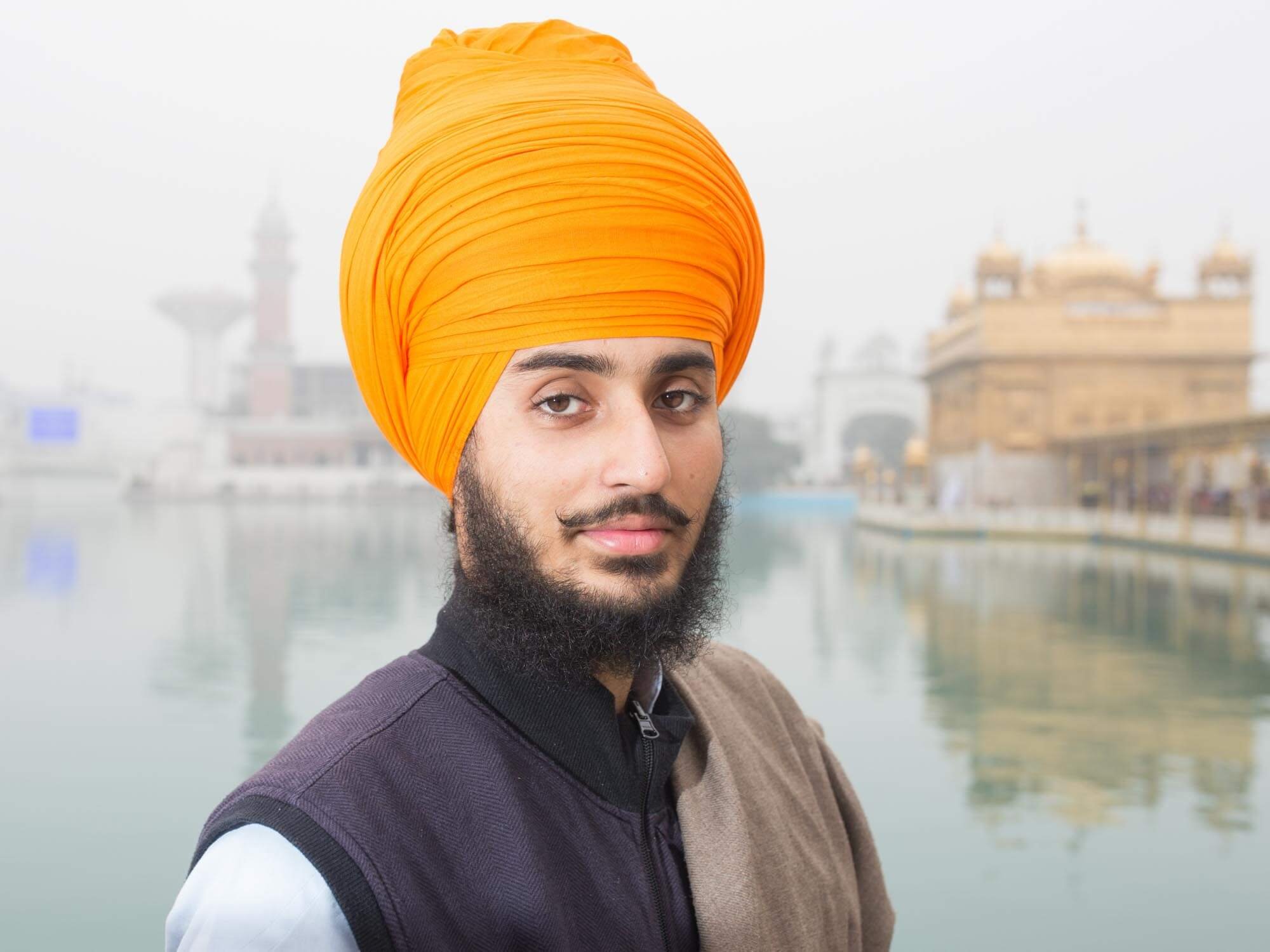 A man wearing an orange turban and traditional Indian clothing standing near a body of water with historical buildings in the background.