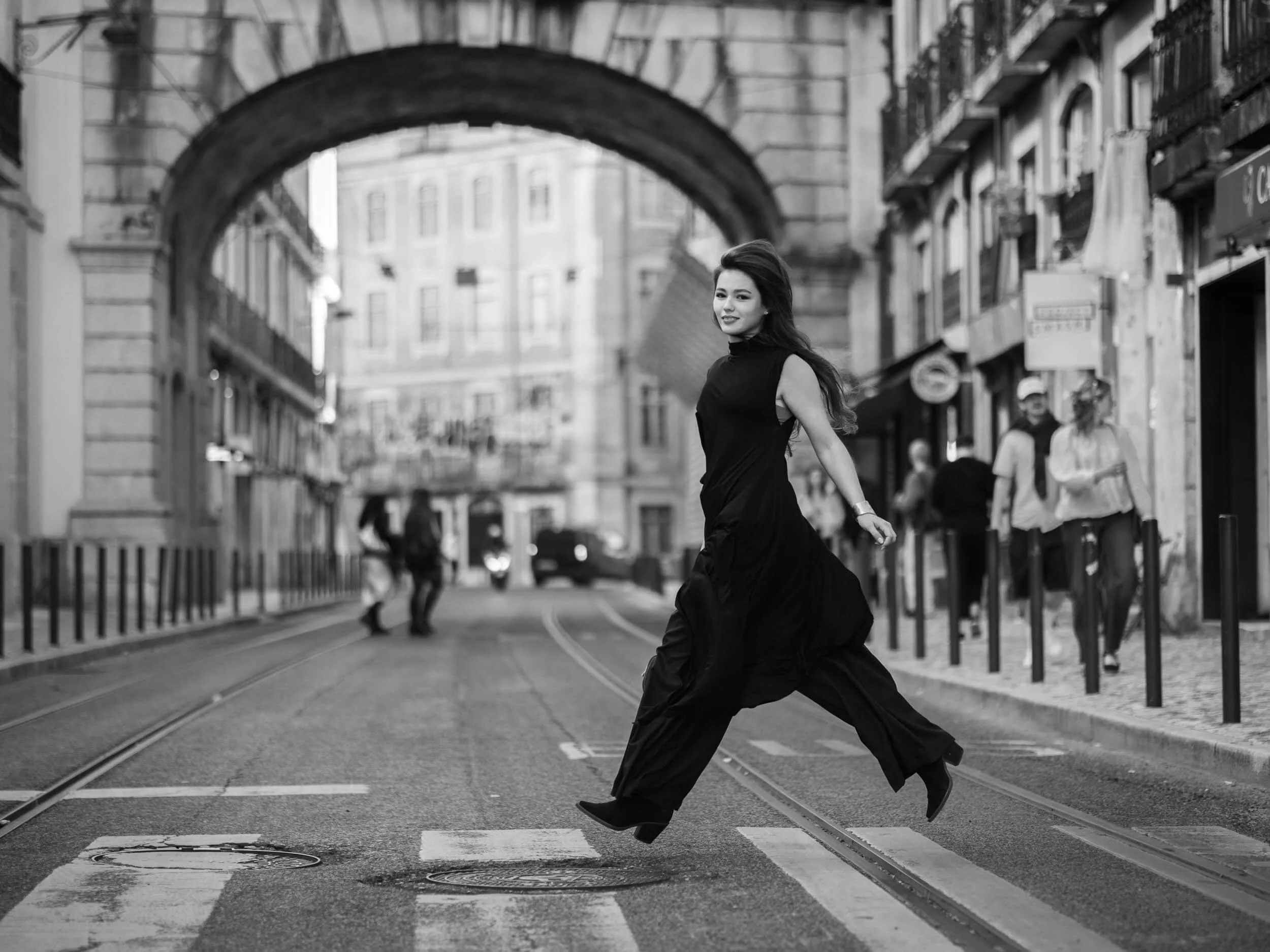 A woman in a black dress crossing a city street with tram tracks, buildings, and pedestrians in the background, captured in black and white.
