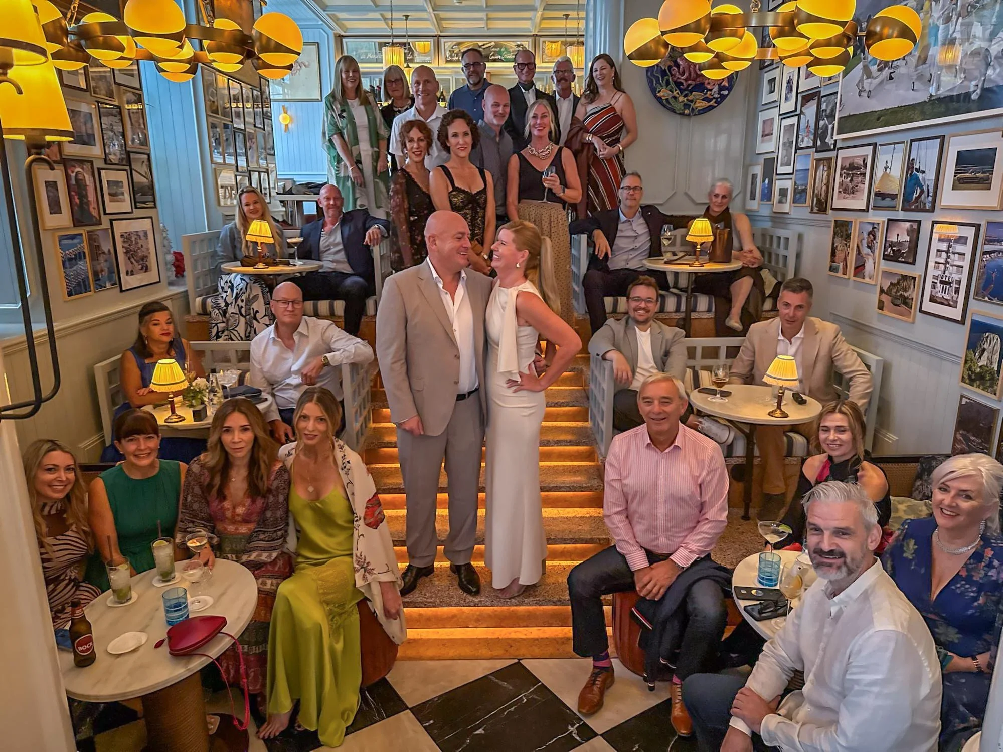 A large group of people gathered in a warmly lit restaurant, posing for a group photo, with two people in the center smiling at each other. The restaurant has framed pictures on the walls and hanging yellow hanging lights.