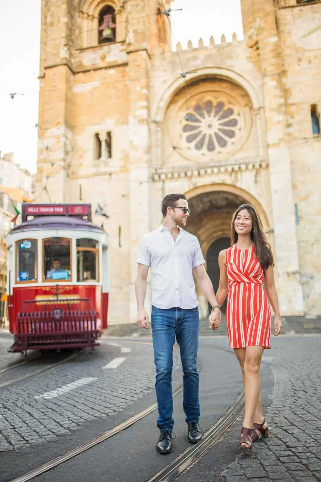A smiling couple posing for the phtoshoot. holding hands walking down a street with a red tram behind them and a Lisbon Cathedral in the Background,