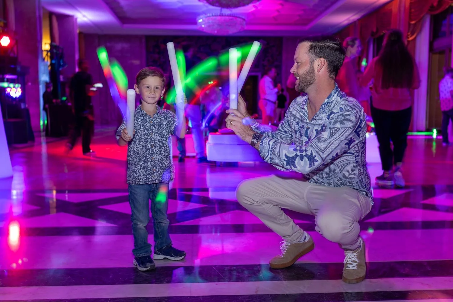 Corporate party at Ritz hotel. A child with a patterned shirt and jeans holding glow sticks, dancing with a man in a patterned shirt and beige pants, crouching and holding a glow stick, in a party setting with colorful lighting and other people in th