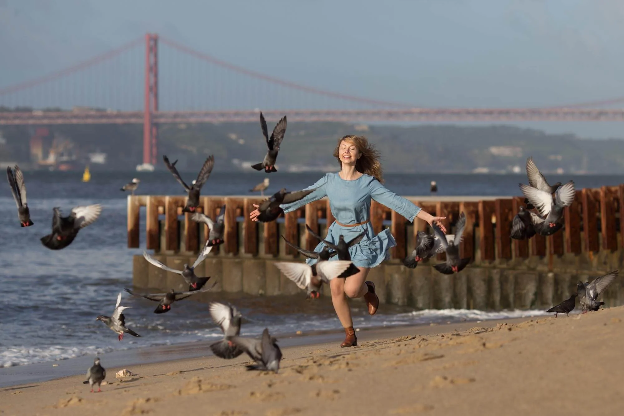 A woman in a blue dress joyfully running along the beach, surrounded by flying pigeons, with a bridge in the background.