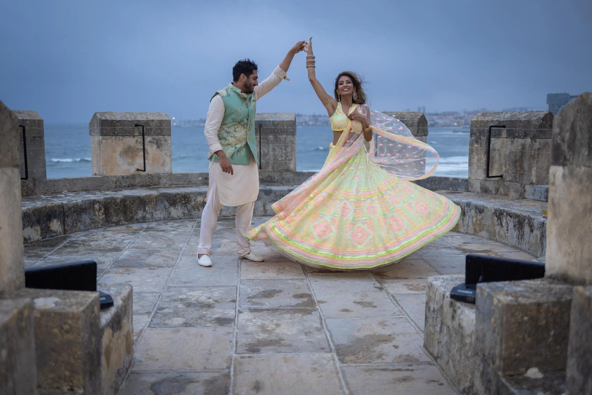 A couple dancing on a stone terrace by the ocean, with a woman in a colorful flowy traditional dress spinning and a man in traditional attire holding her hand.