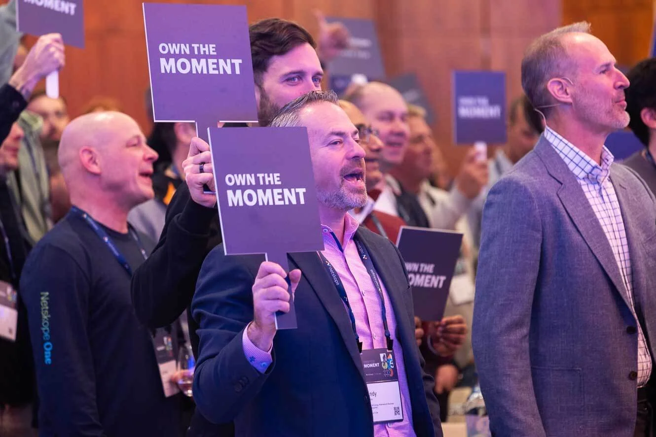 A group of people at a conference holding purple signs that read 'OWN THE MOMENT' during a presentation or applause.