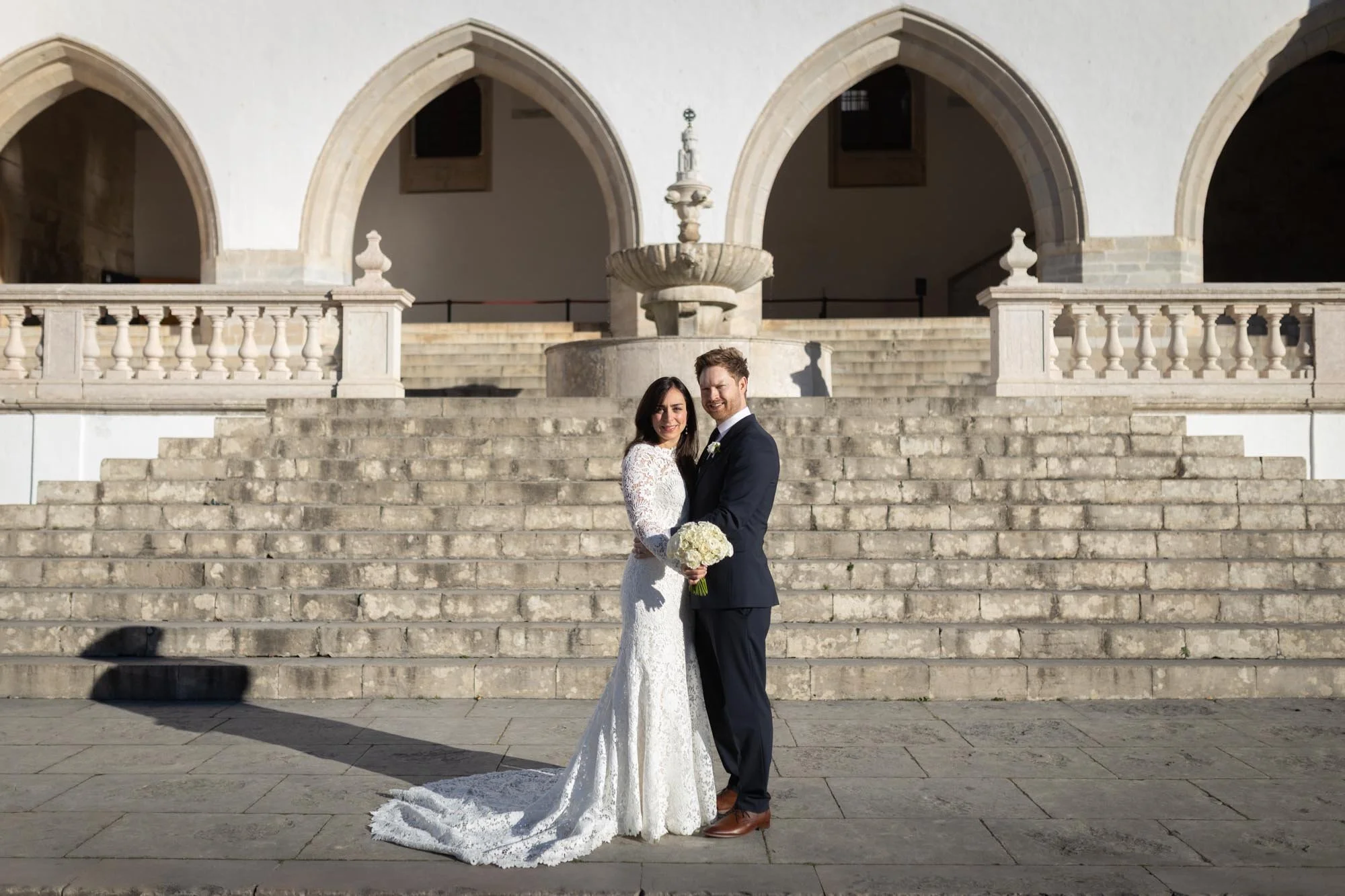 A bride and groom standing on stone steps in front of an ornate fountain and arched building, celebrating their wedding day.