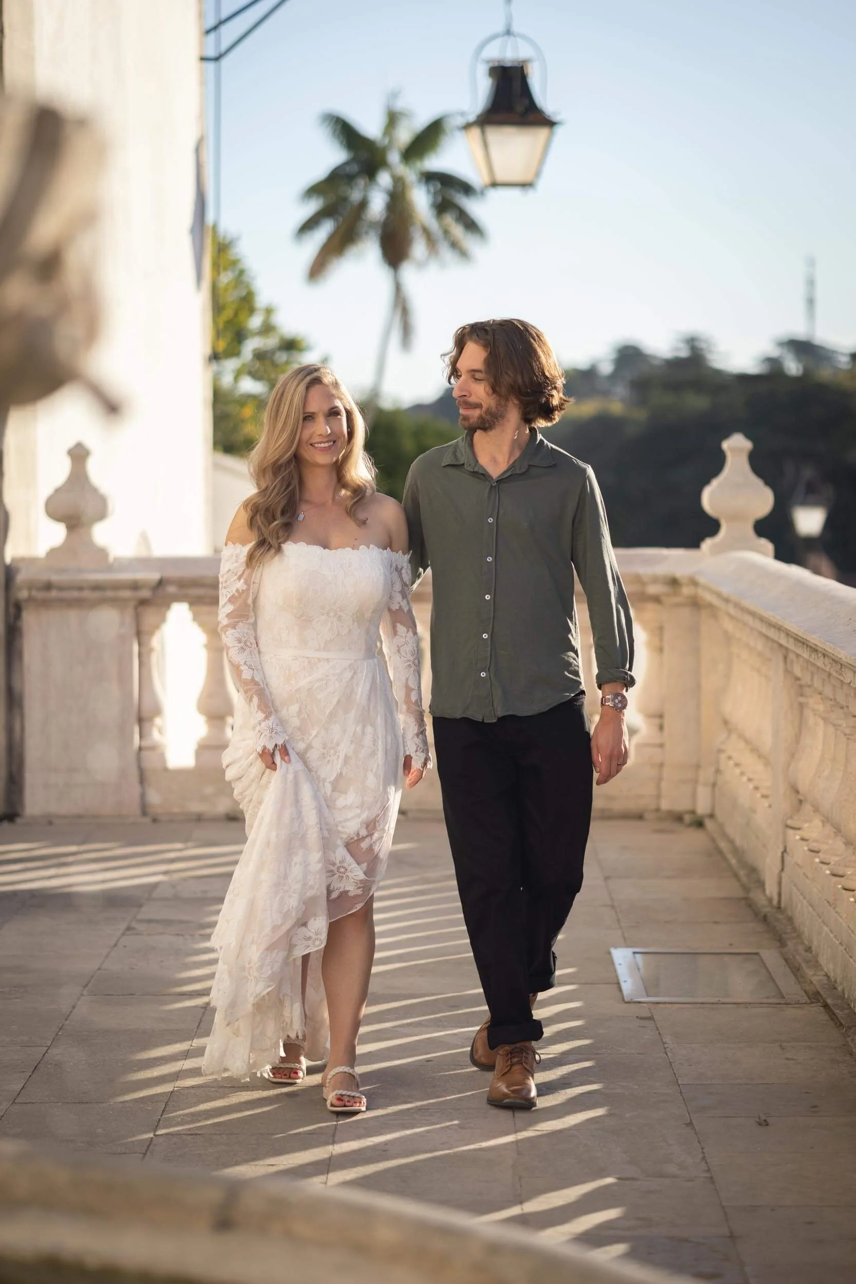 A woman in a white lace dress and a man in a dark green shirt and black pants walking together on a stone pathway with a balustrade, lantern, palm trees, and blue sky in the background, during a engagement photoshoot in Sintra.
