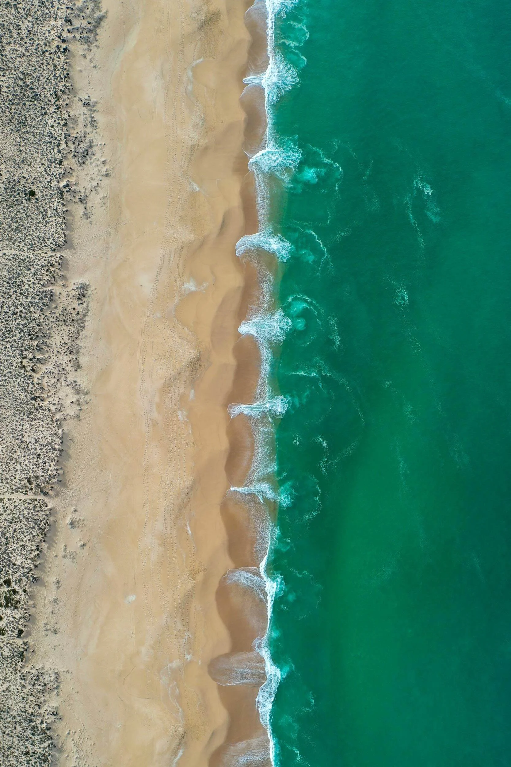 Aerial view of a sandy beach with green ocean waves crashing onto the shore, with some sparse vegetation on the left side.