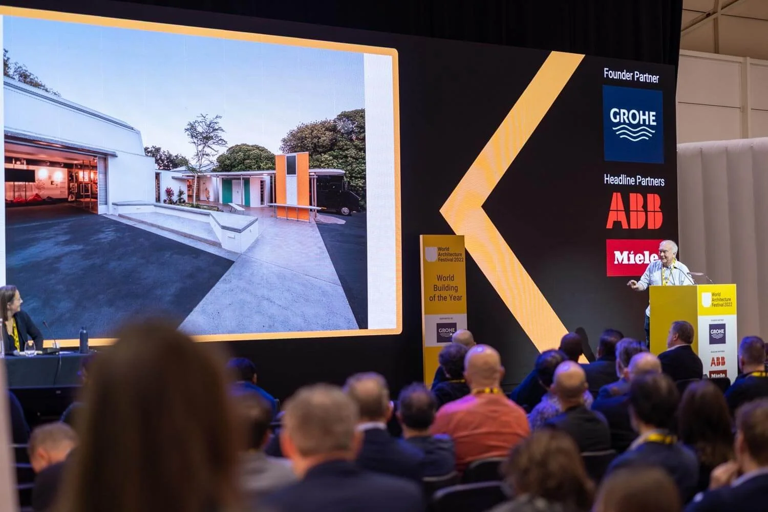 A presentation at an architecture event showing a large screen with an outdoor architectural design, and an audience watching the speaker on stage. The event is titled 'World Architecture Festival 2022' and 'World Building of the Year'.