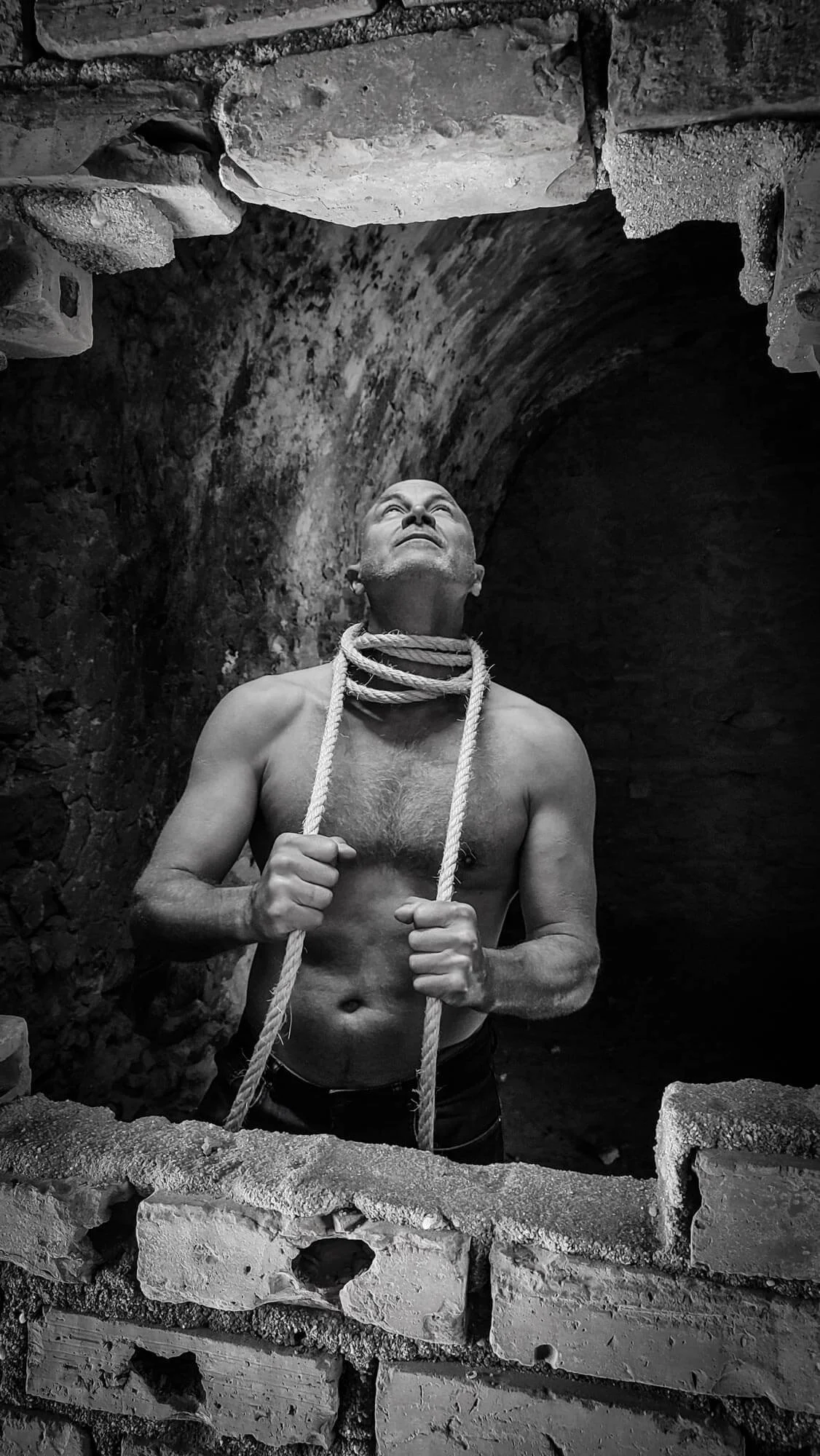 A shirtless man standing in a brick structure, looking up, with a rope around his neck, in a dramatic black-and-white photo.