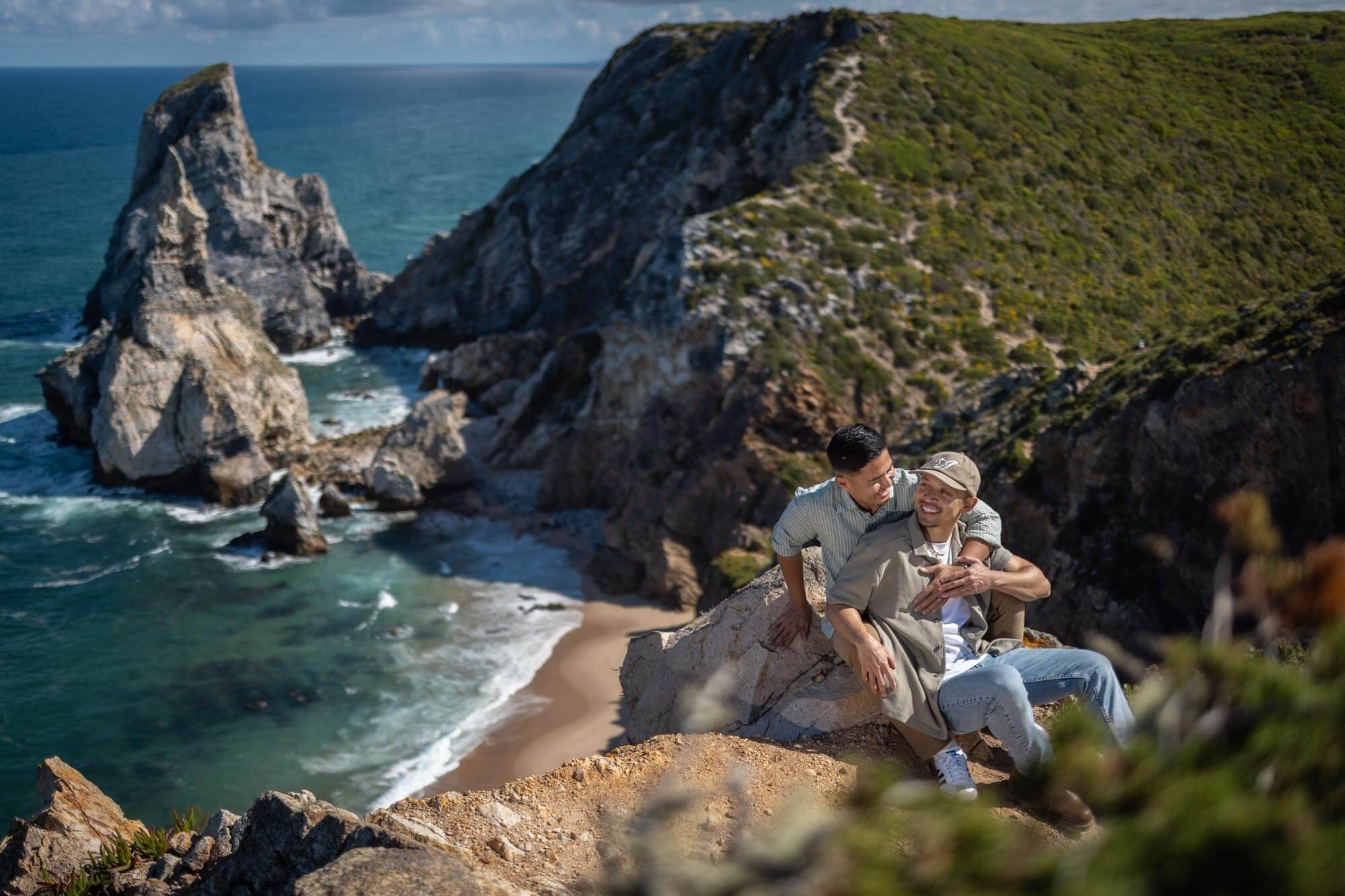 Two men sit on a rocky cliff overlooking a beach and ocean, smiling and enjoying each other's company with cliffs and greenery in the background.