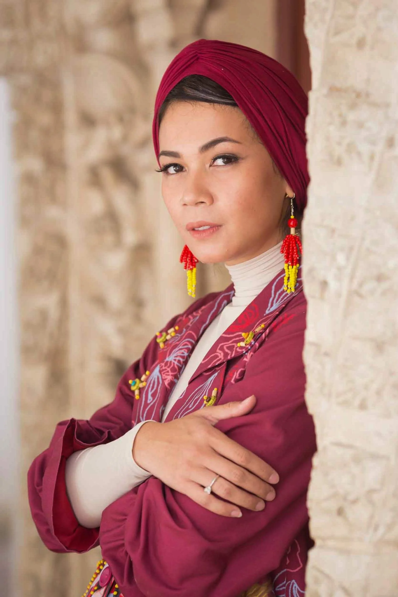A woman with medium skin tone wearing a red headscarf, red and yellow beaded earrings, and a maroon jacket with embroidered designs, standing against a stone wall.