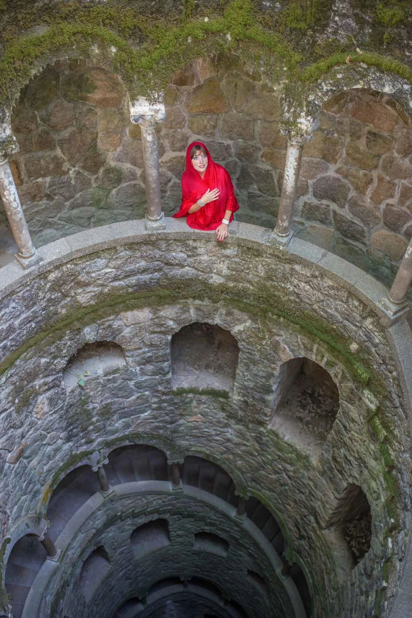 A woman with red hair wearing a red shawl and jewelry, standing at the top of a stone well or tower with moss-covered stone walls and columns, looking up towards the camera.