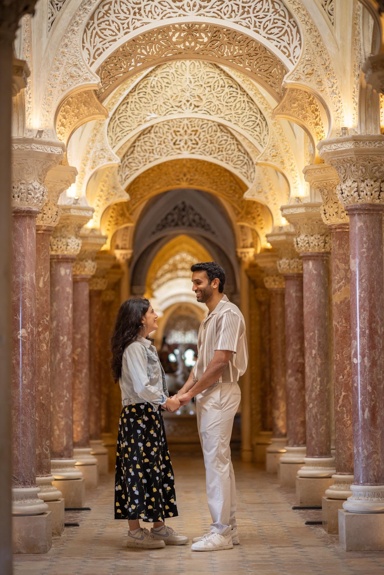 A couple holding hands and smiling at each other in an ornate, beautifully lit corridor with intricate architectural details, arches, and columns.