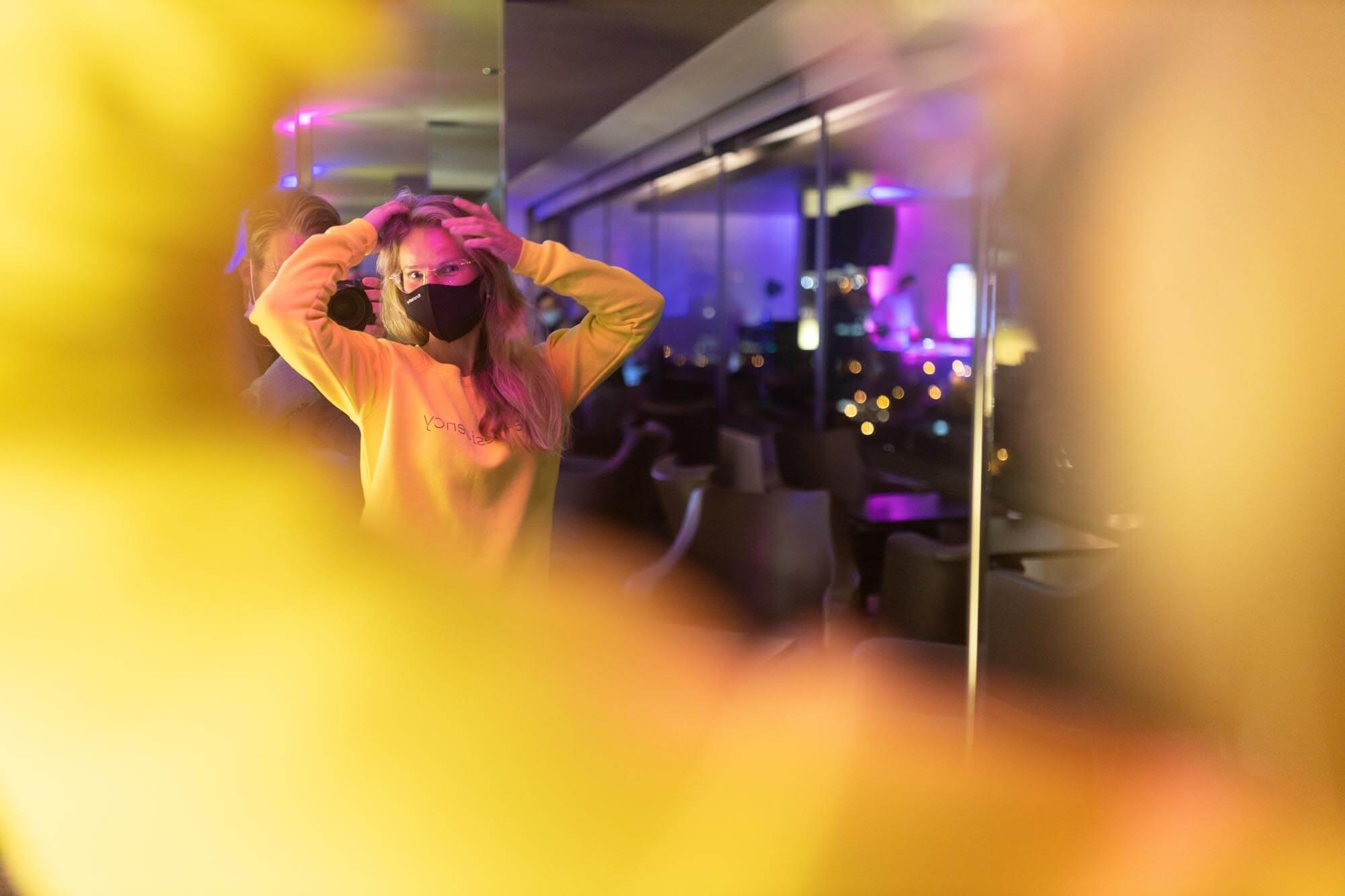 A woman wearing glasses and a black face mask, adjusting her hair, is taking a mirror selfie in a dimly lit space with purple and yellow lighting.