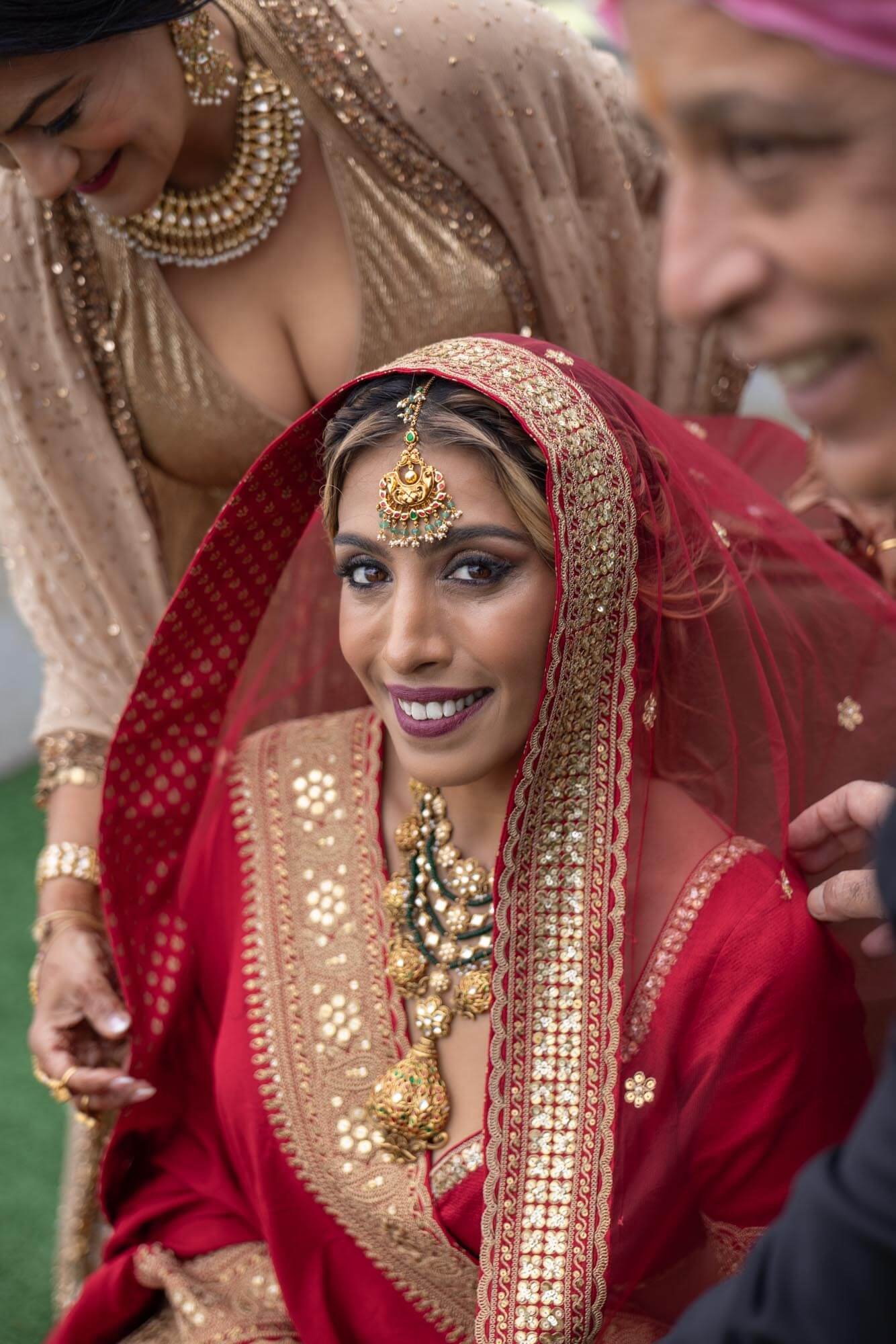 A bride dressed in traditional red and gold Indian wedding attire with intricate jewelry, smiling, surrounded by women also in vibrant traditional dresses.