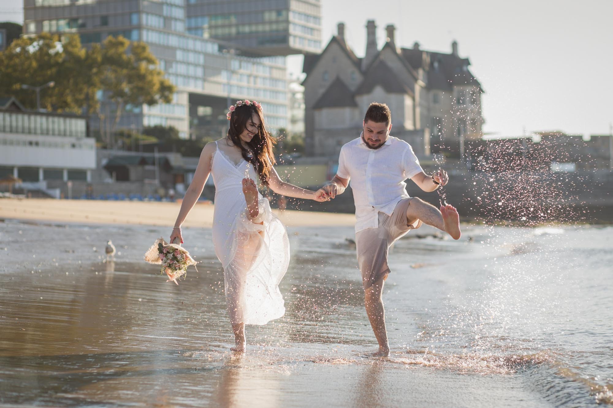A couple holding hands and playing in the water at the beach during sunset, with city buildings and a historic house in the background.