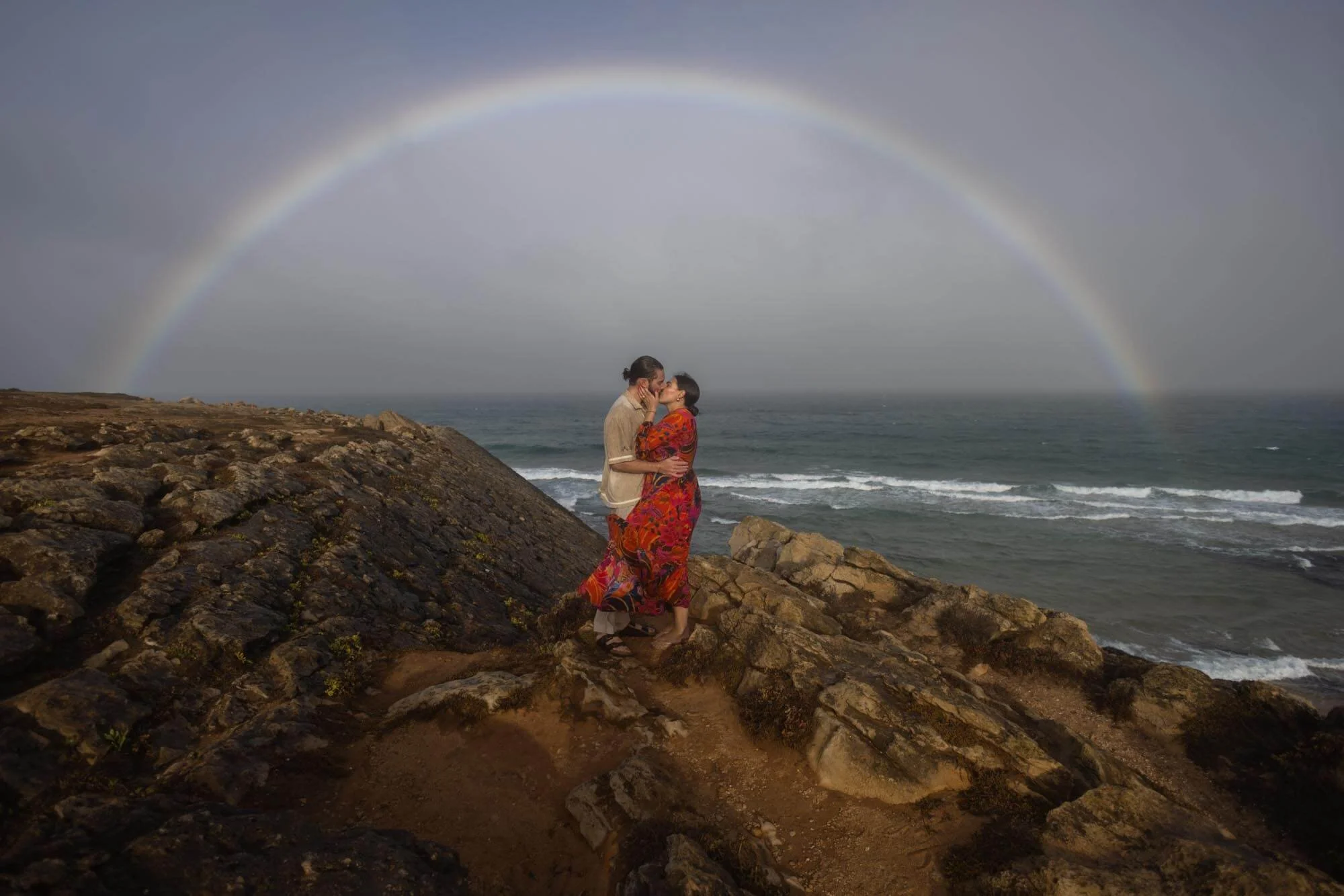 Two women sharing a kiss on a rocky coastline with a rainbow in the background.