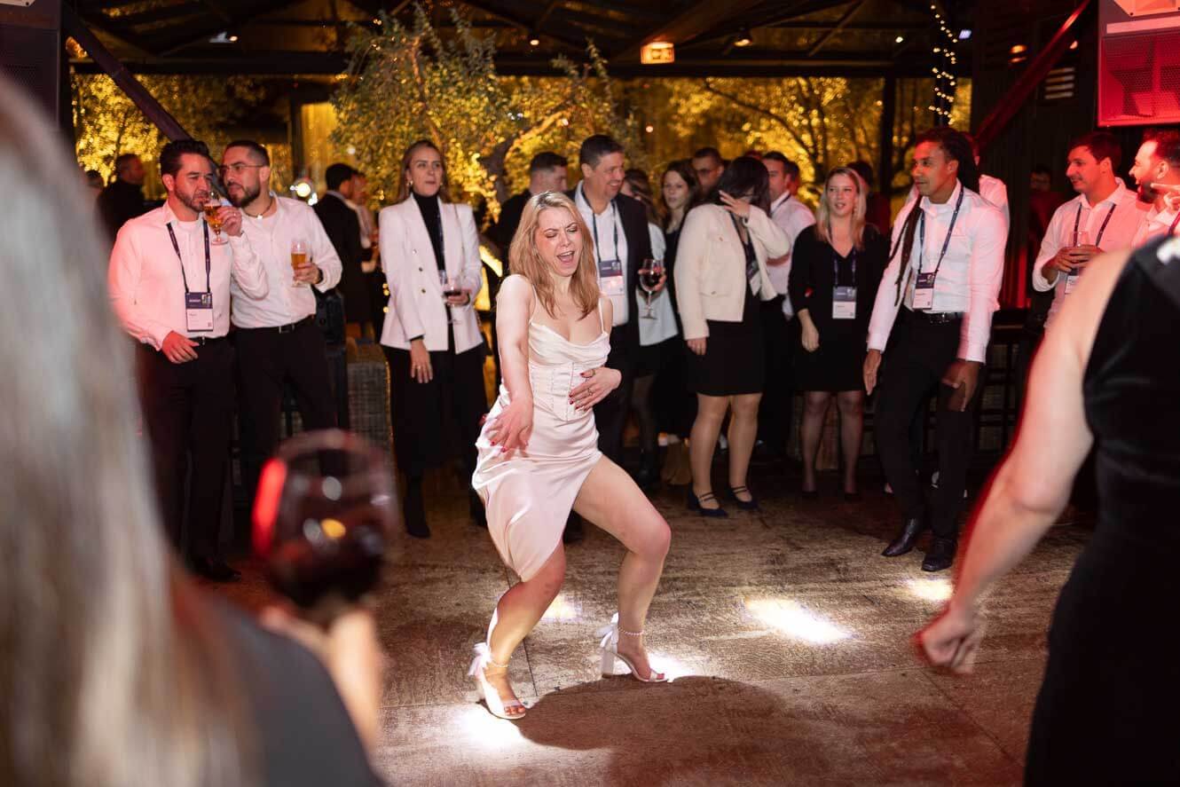 A woman in a white satin dress dancing at a party with guests watching and drinking beer in a warmly lit indoor venue.