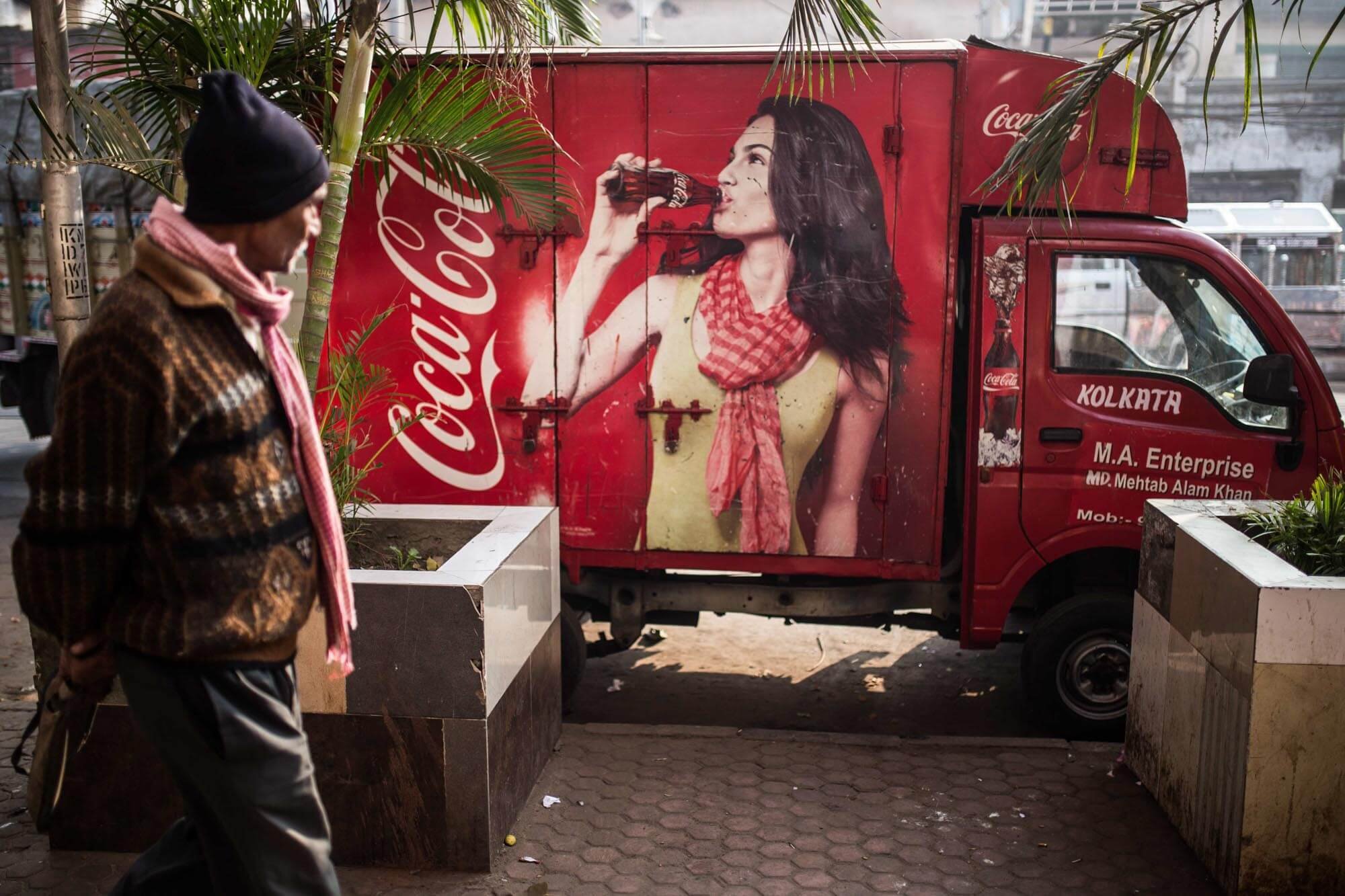 A man walking past a red Coca-Cola delivery truck with a large advertisement of a woman drinking Coca-Cola, situated on a sidewalk with potted plants.