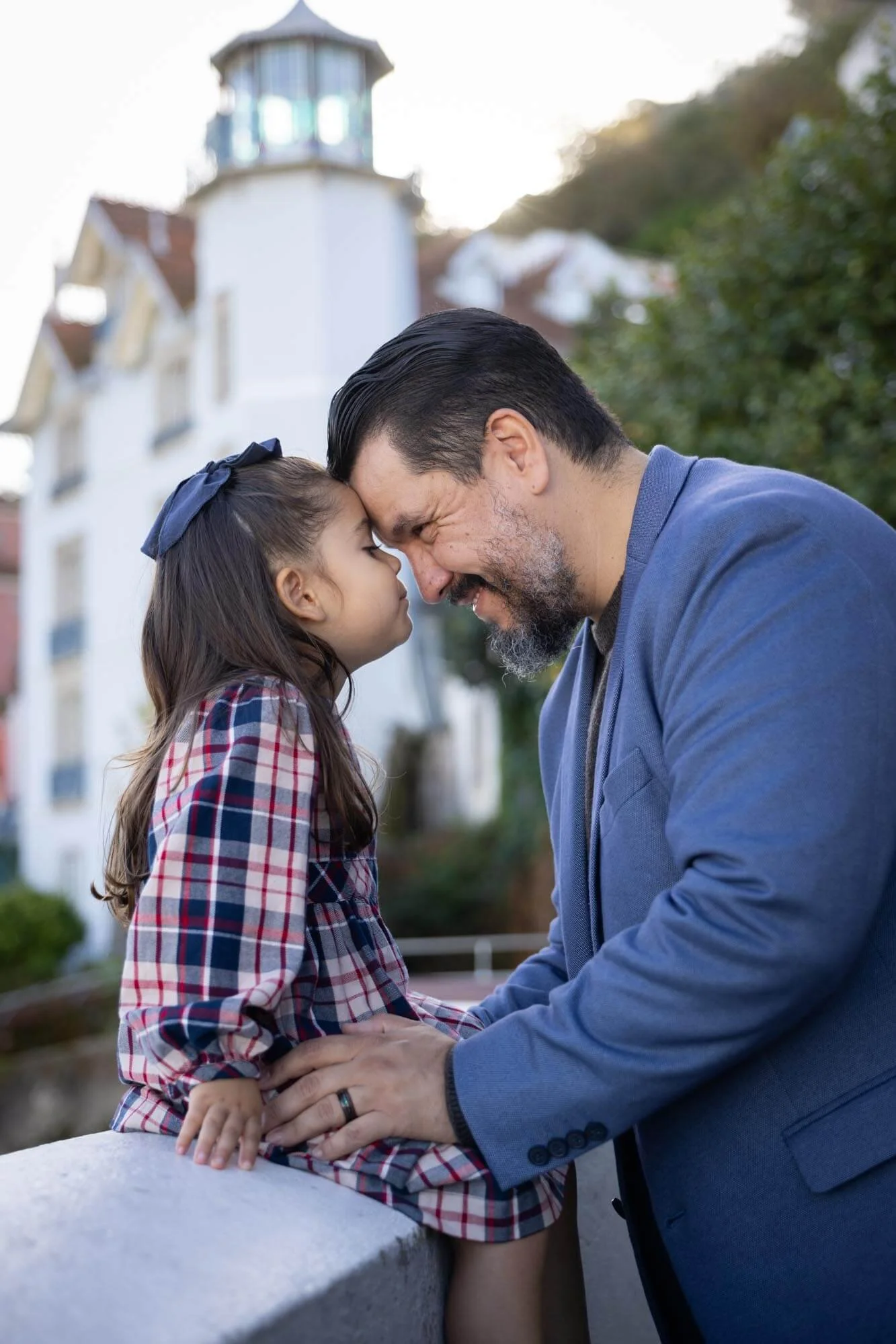A father and daughter with foreheads touching, smiling and enjoying a tender moment outdoors with a lighthouse and a white building in the background.