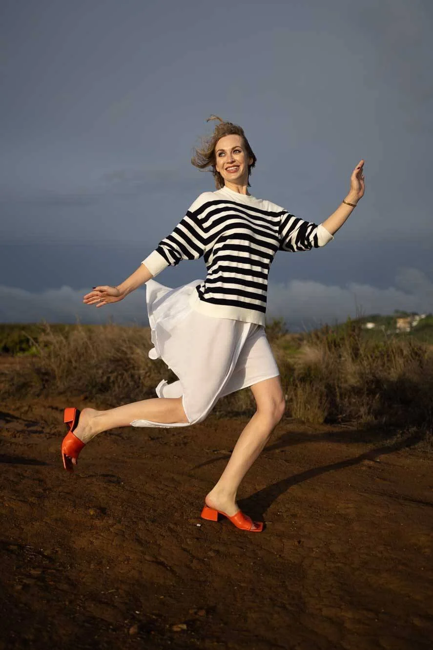 A woman in a striped sweater and a white skirt running outdoors on a dirt path, with clouds in the sky behind her.