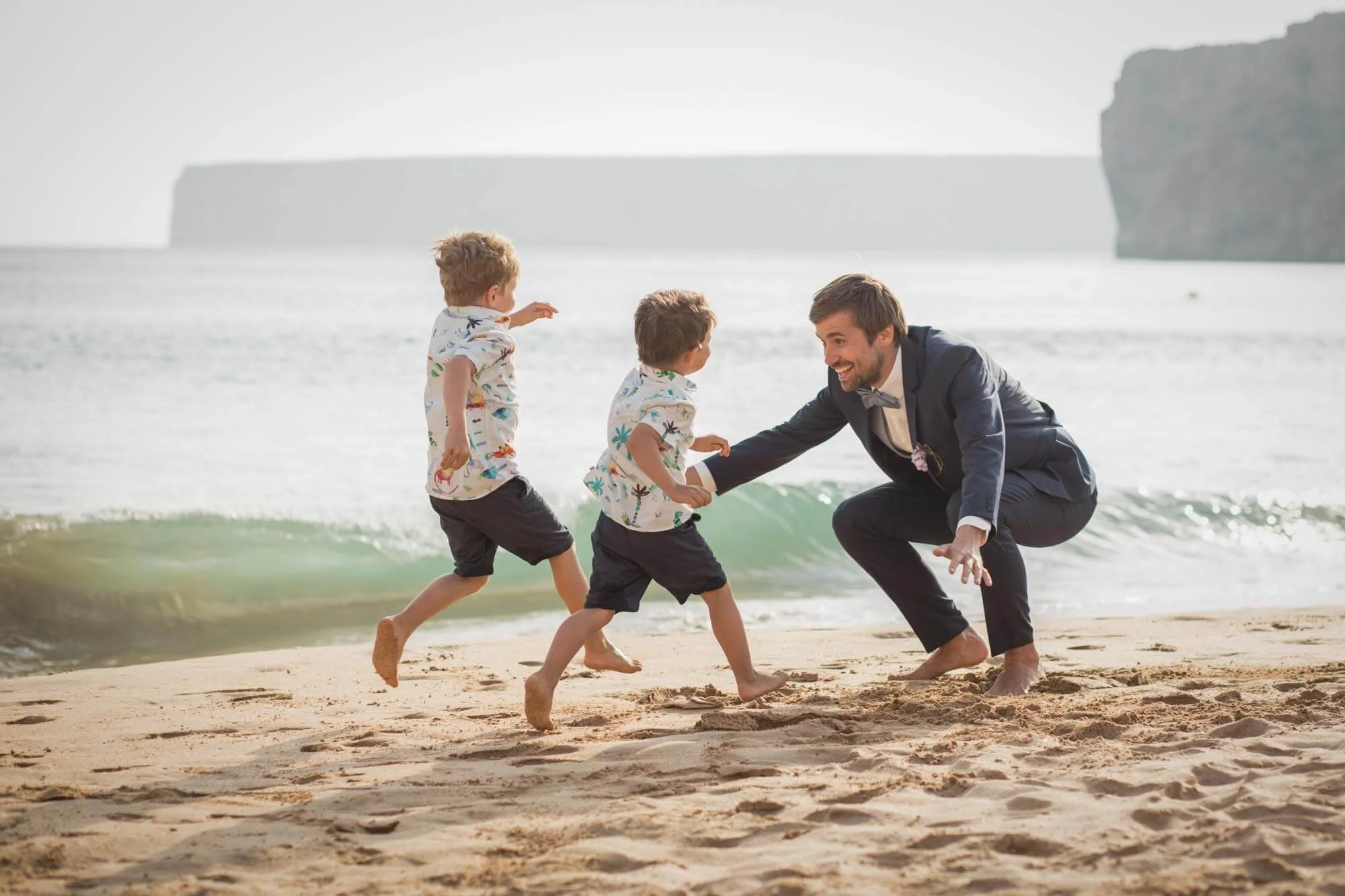 A man and two children playing and running on the beach near the ocean, with cliffs in the background.